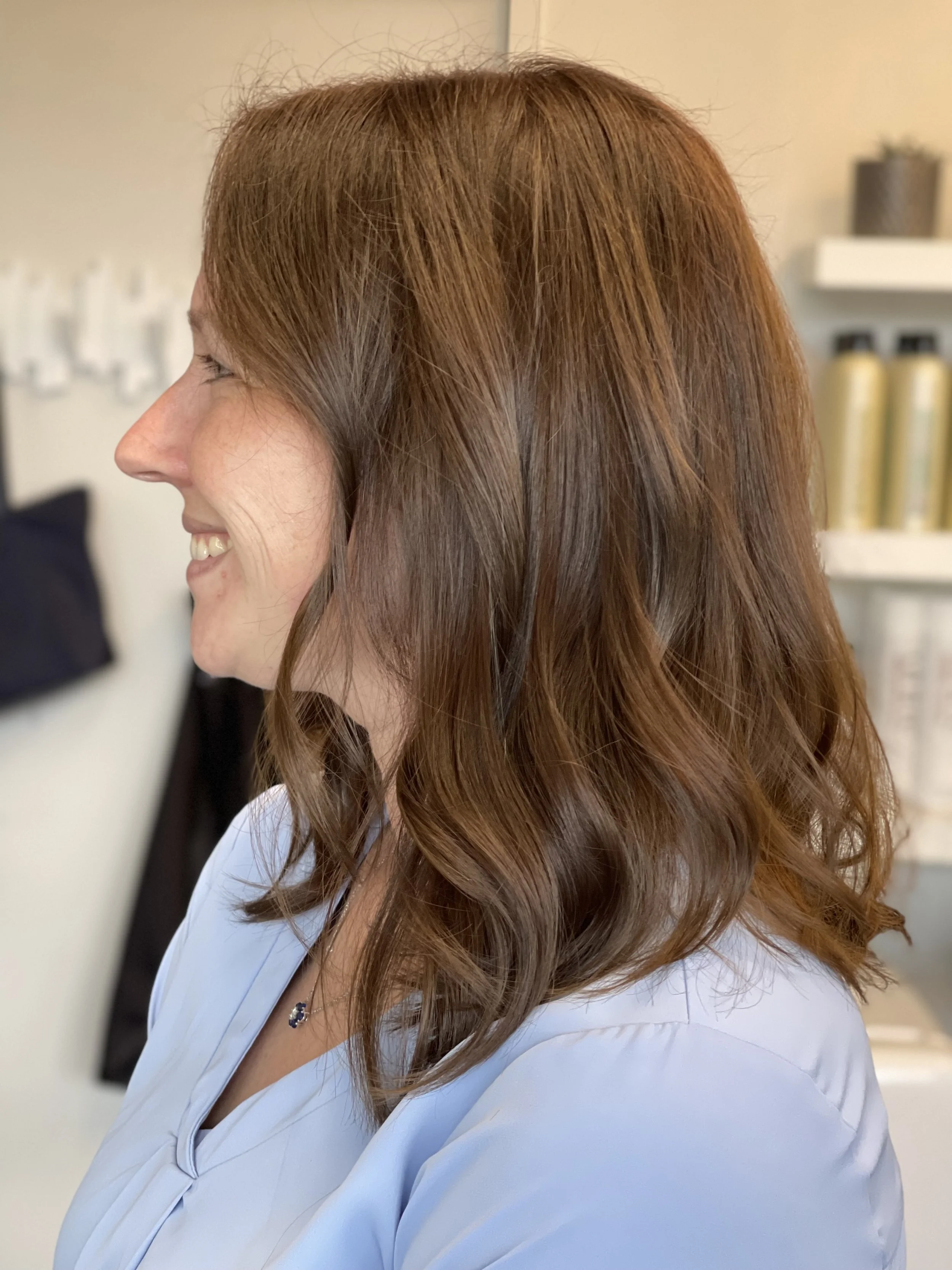 A woman with shoulder-length wavy red hair wearing a light blue shirt, smiling in a salon or similar setting.