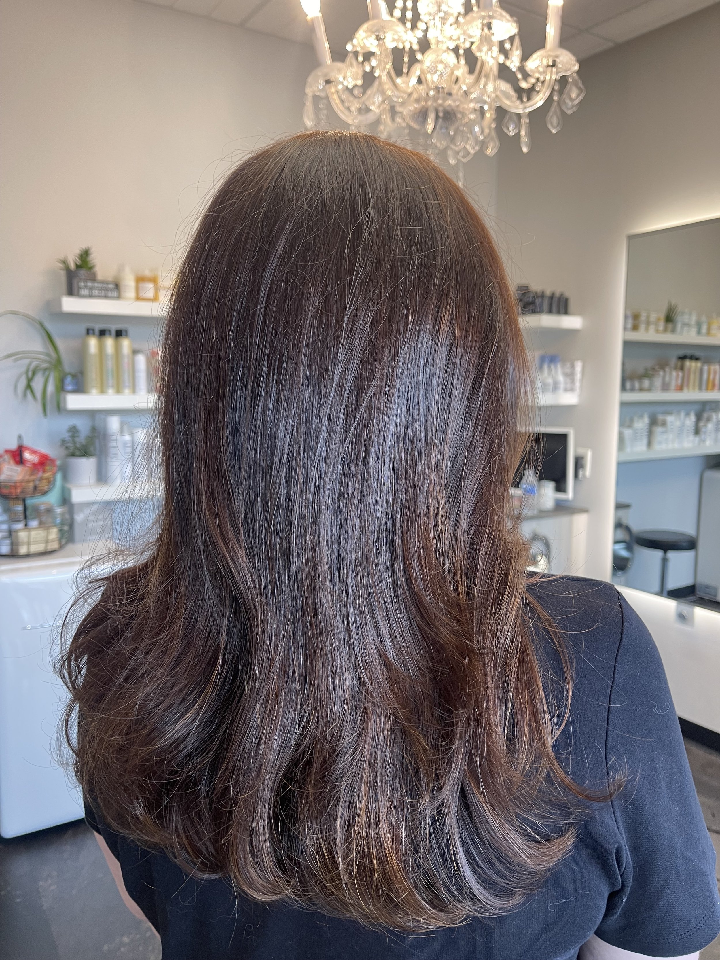 Back view of a woman with mid-length brown hair with subtle highlights, in a salon with shelves of hair products and a chandelier in the background.