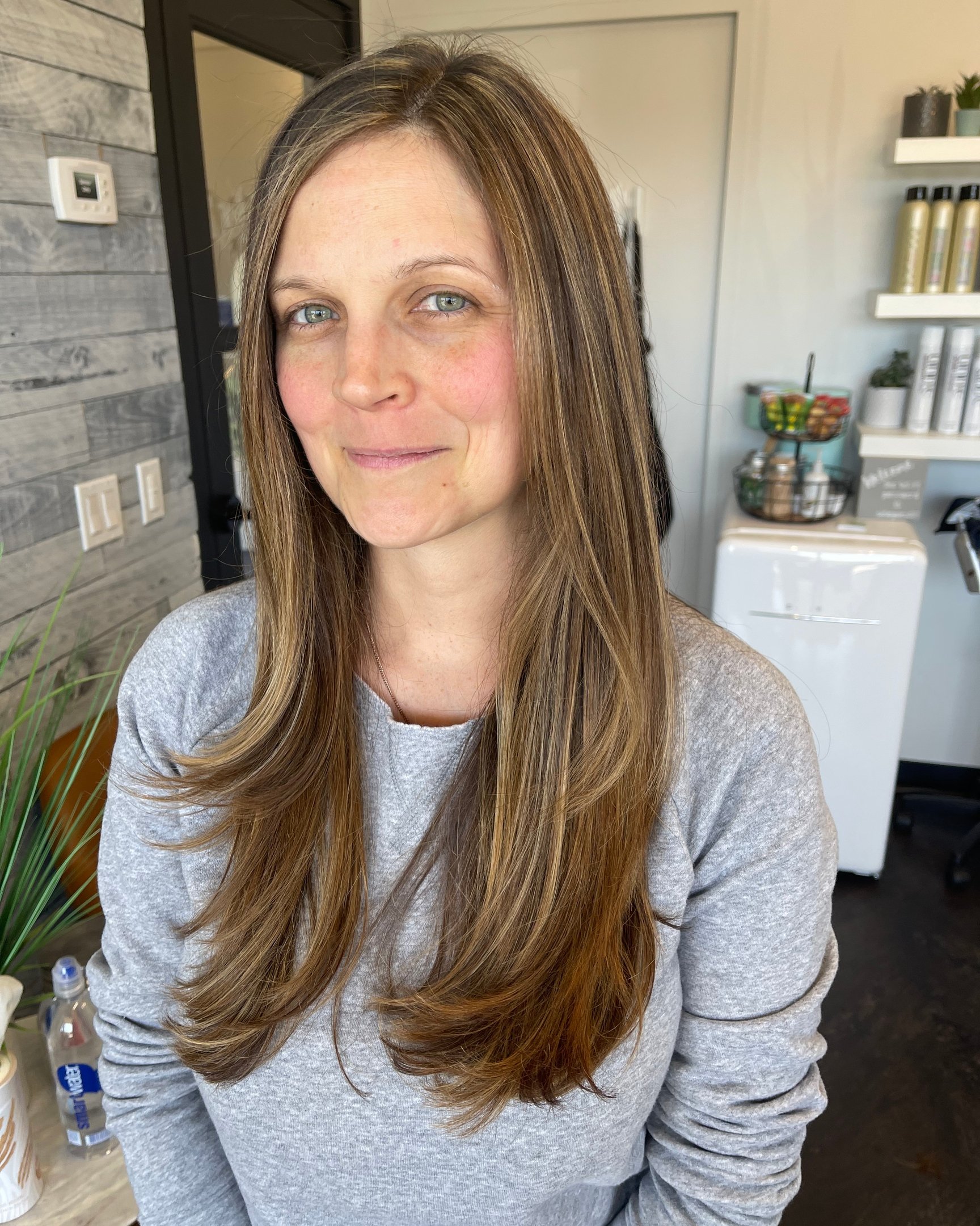 A woman with long light brown hair and blue eyes, wearing a gray sweatshirt, smiling softly in a cafe or coffee shop setting with plants and shelves in the background.