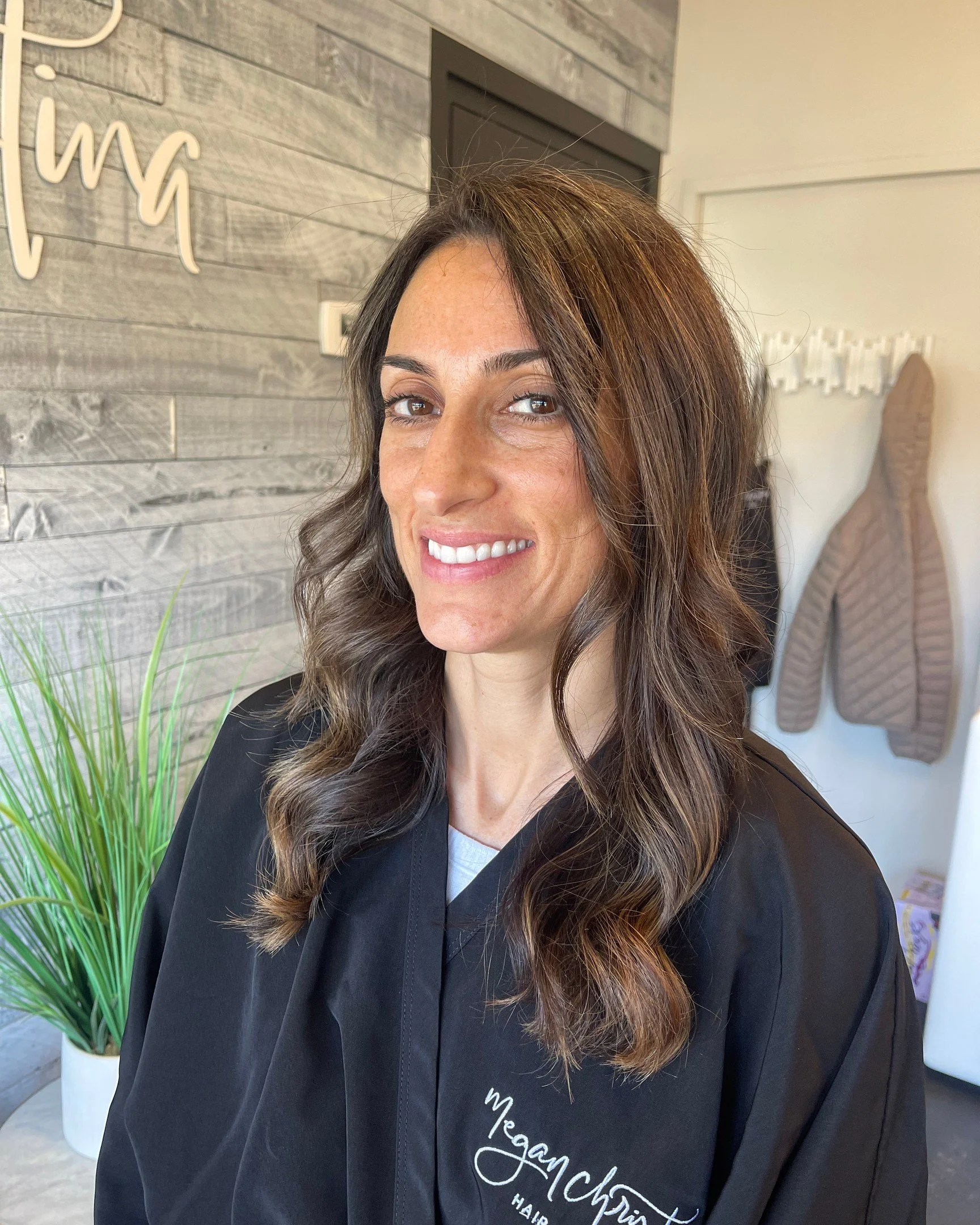 A woman with long wavy brown hair and light skin smiling at the camera. She is wearing a black salon cape with the words 'Megan McInt' embroidered on it. The background features a light gray wooden wall, a potted green plant, and a brown quilted jack