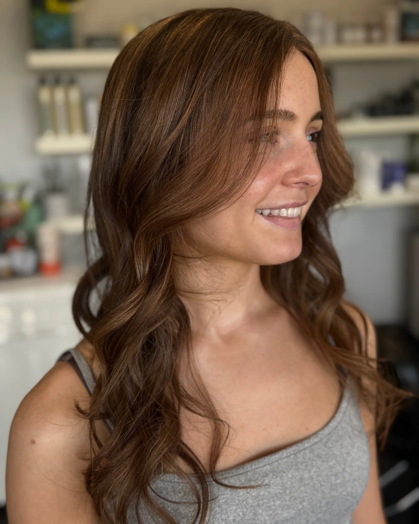A woman with long, wavy auburn hair smiling in a kitchen.