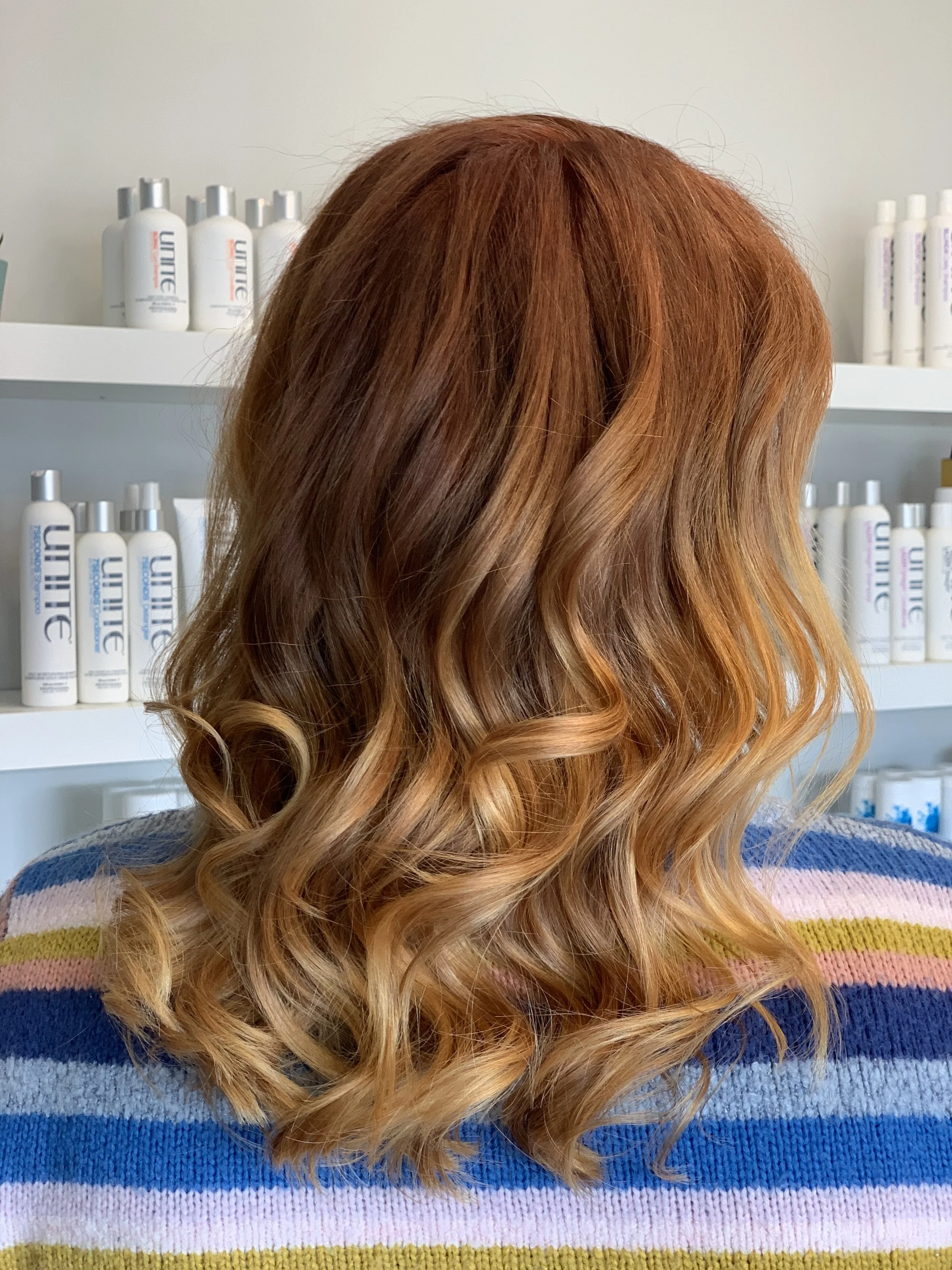 Back of a woman with wavy, reddish-brown hair wearing a colorful striped sweater, standing in front of shelves with hair care products.