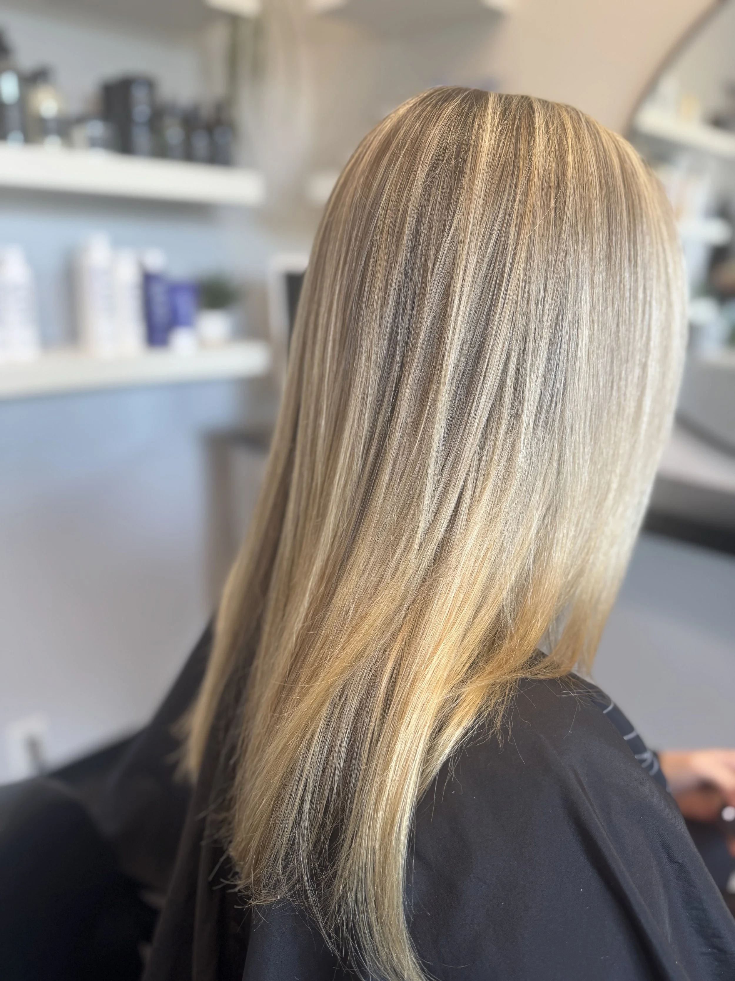 Back of a woman's head showing her straight blonde hair with highlights, sitting in a salon or barber shop.