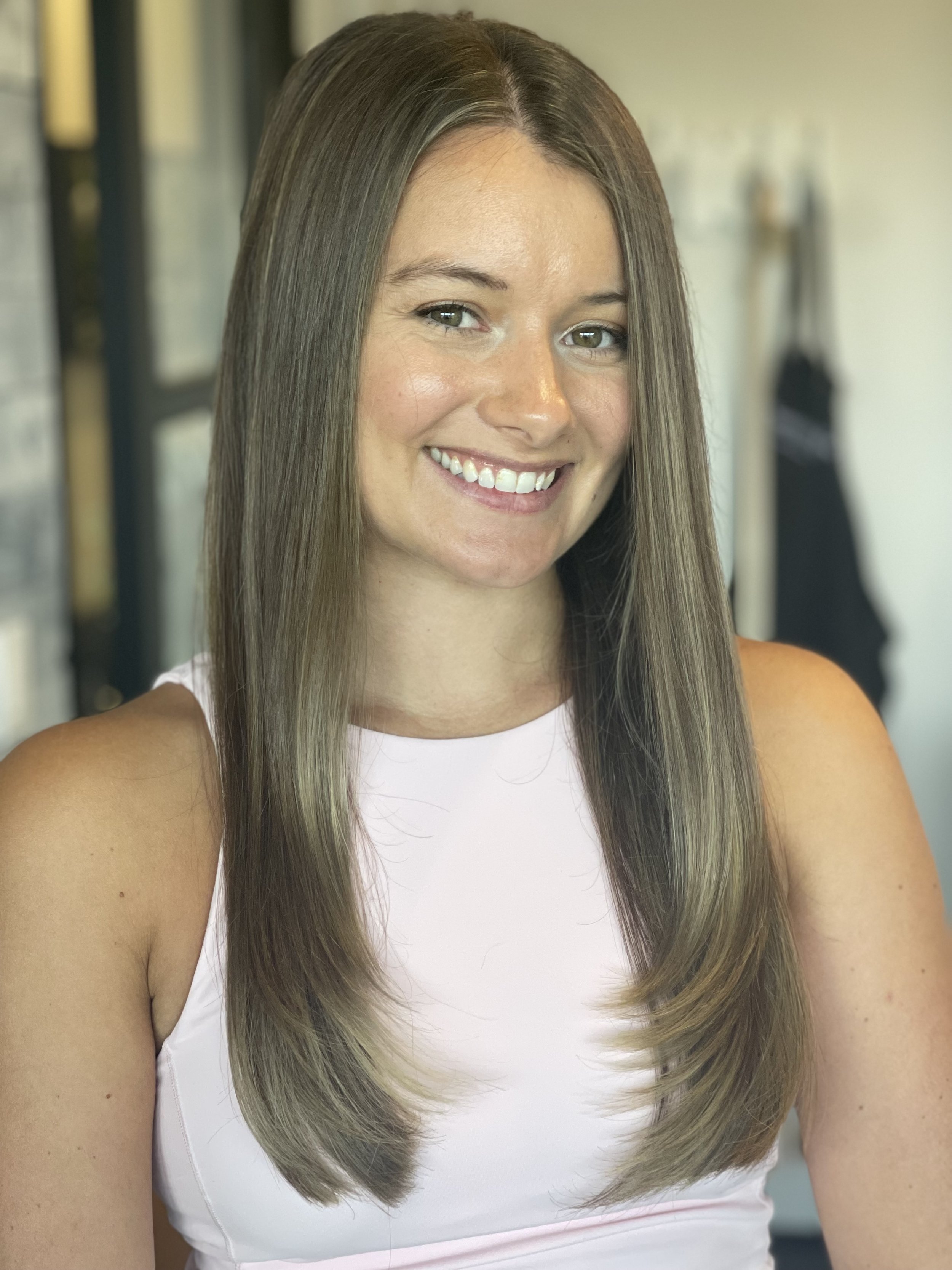 A woman with long, straight brown hair, smiling and wearing a sleeveless light pink top.