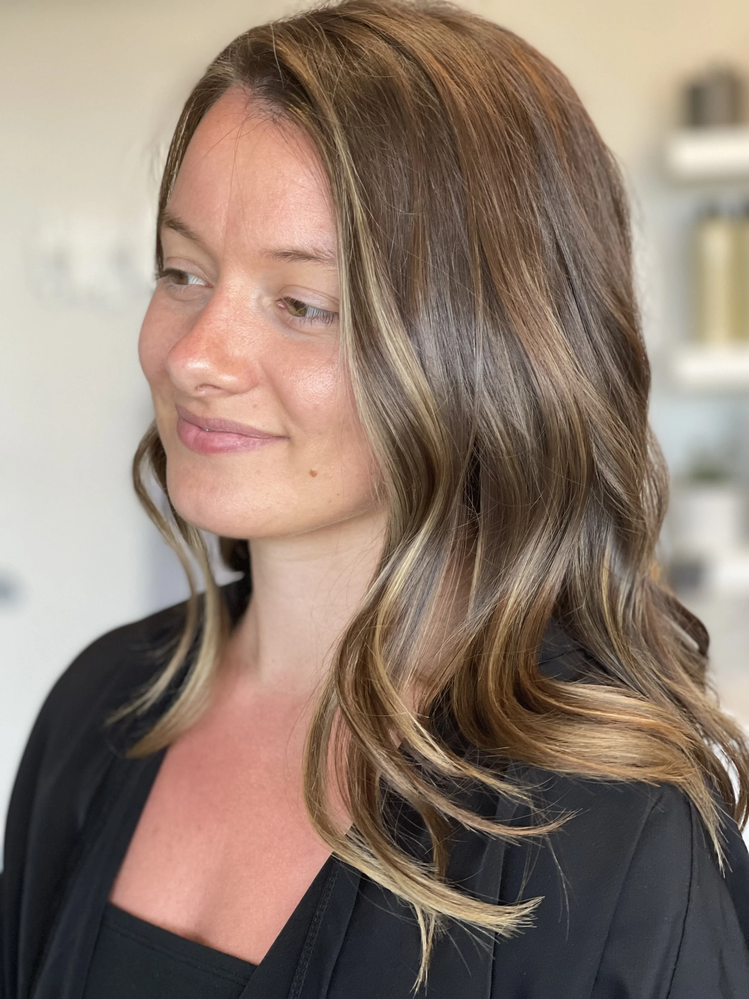A woman with wavy, shoulder-length brown hair with blonde highlights, wearing a black top, smiling softly with eyes cast downward.