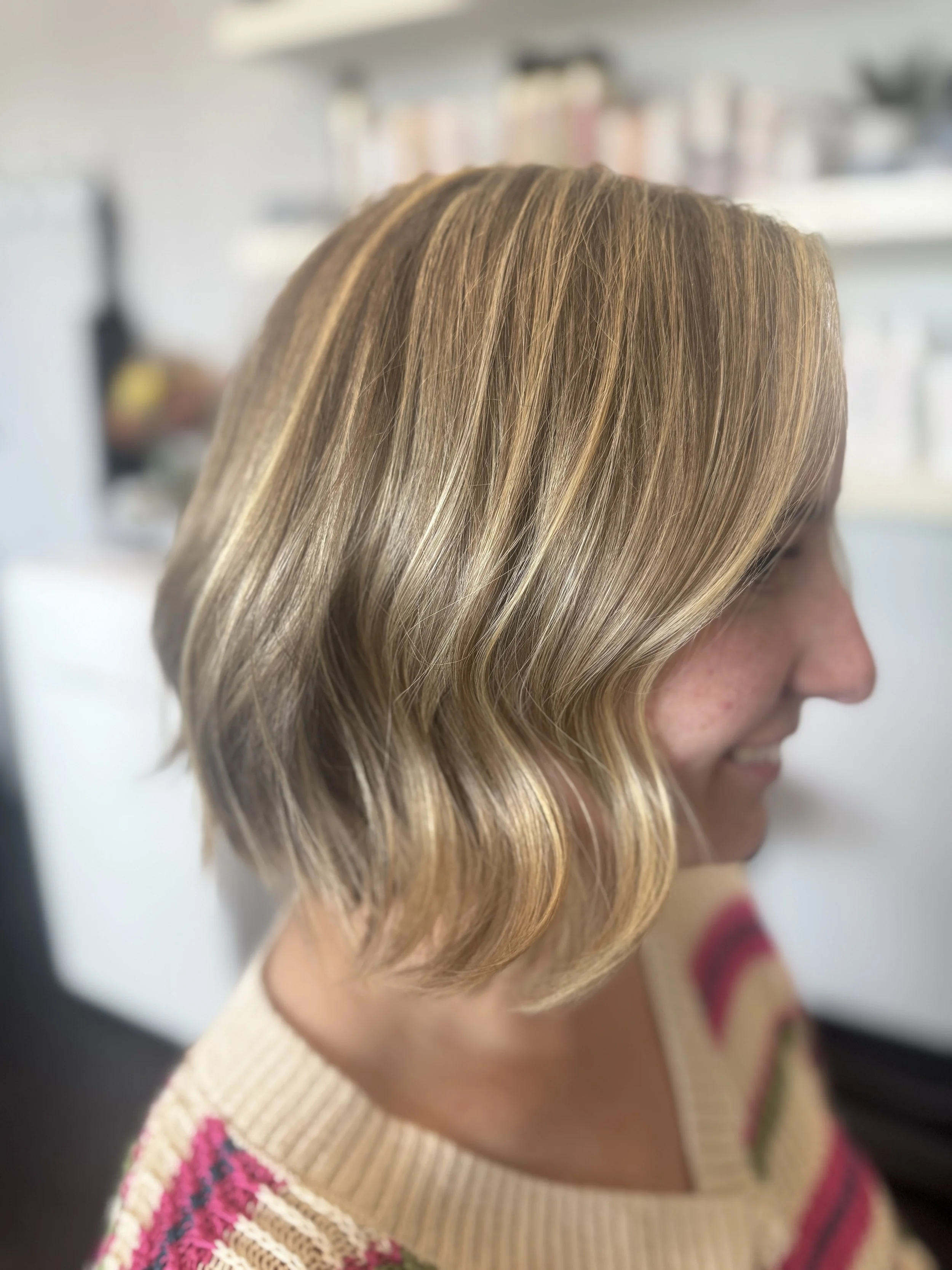 Side view of a woman with shoulder-length blonde hair styled in loose waves, wearing a colorful sweater with pink, beige, and green stripes, smiling.