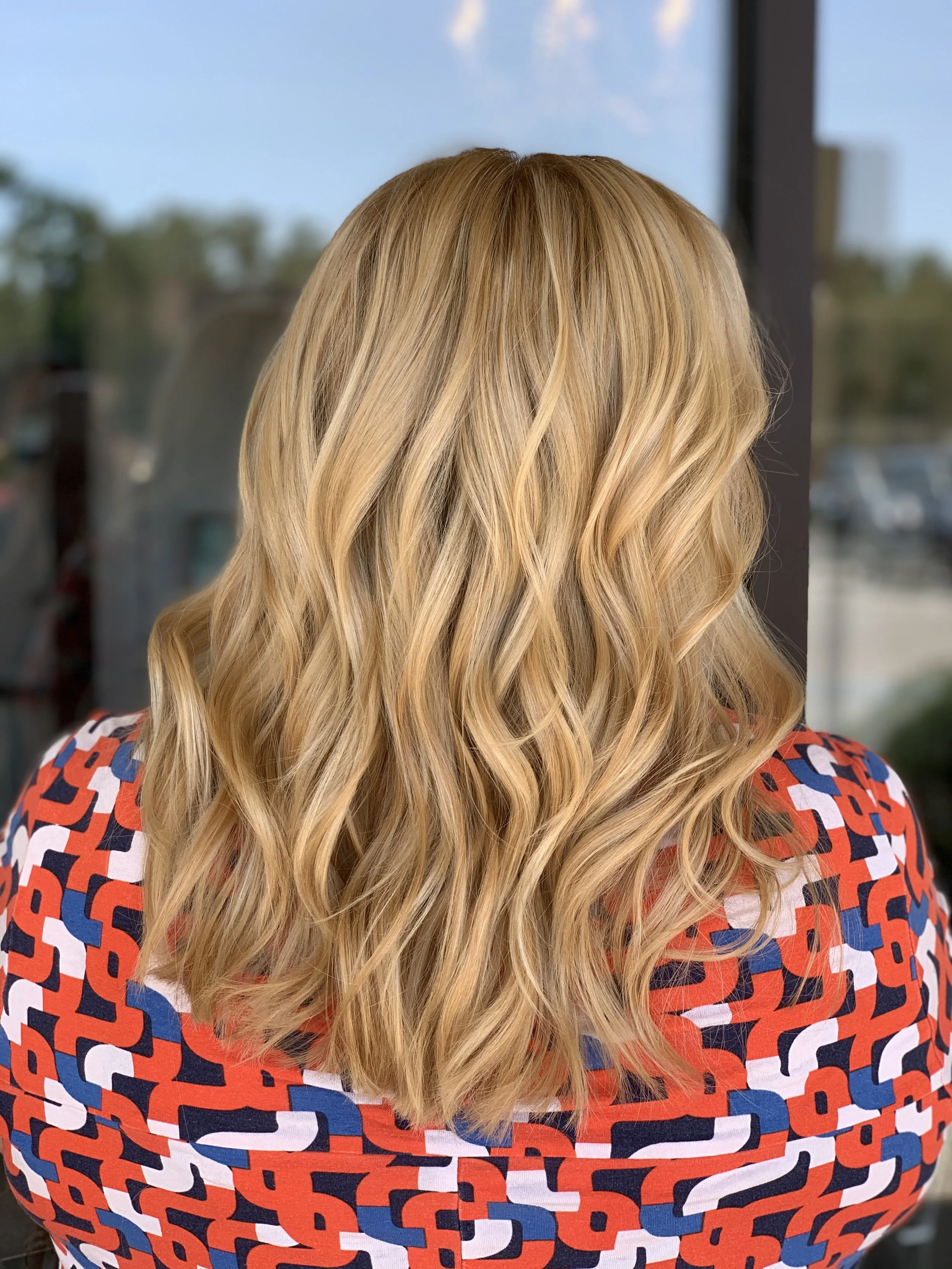 Back of a woman with long, wavy blonde hair wearing a multicolored geometric patterned top, standing indoors but near a window with a view of blue sky and trees outside.