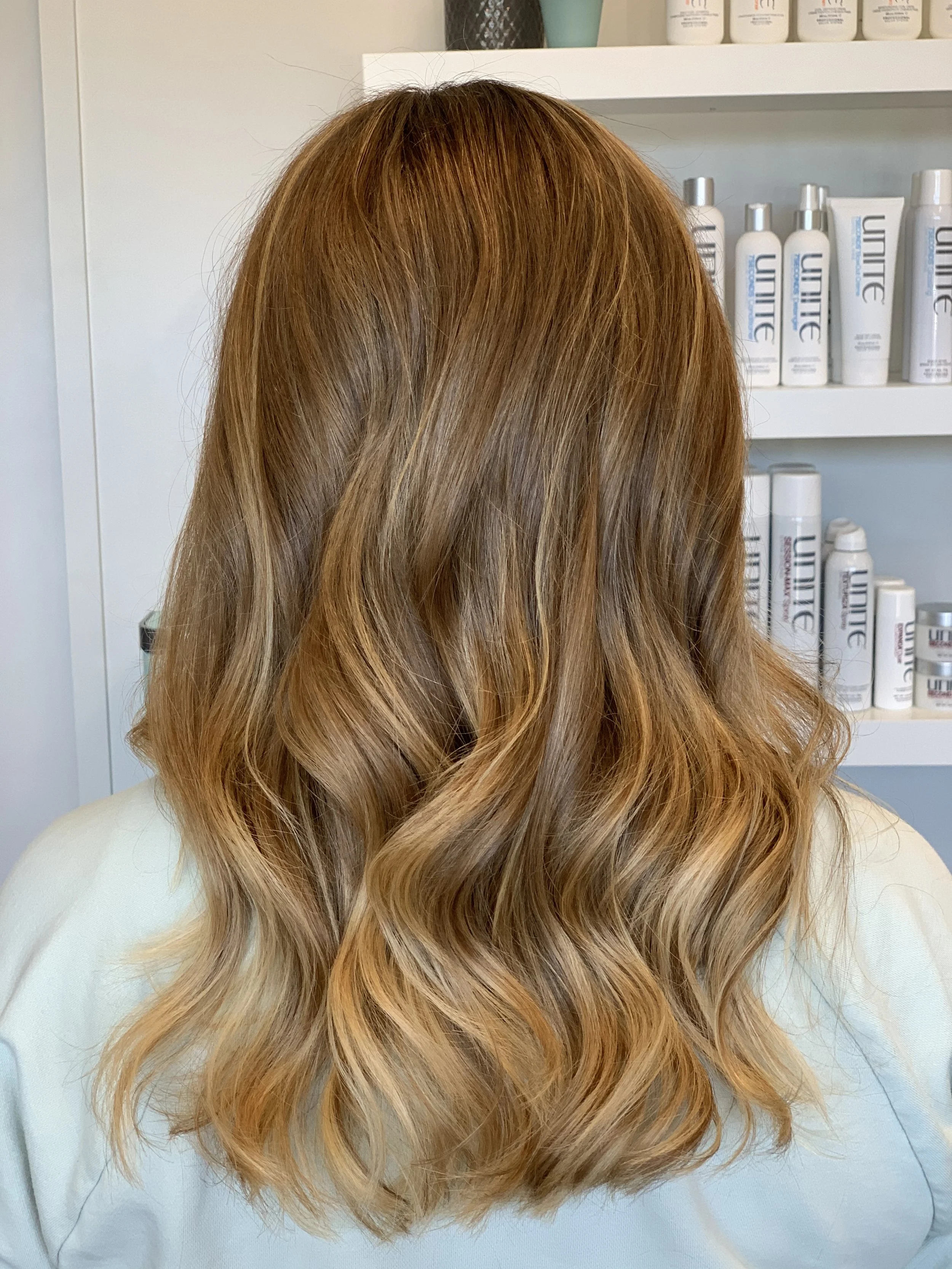 Back of a person's head with long, wavy, reddish-brown hair, in a hair salon with shelves of hair products in the background.