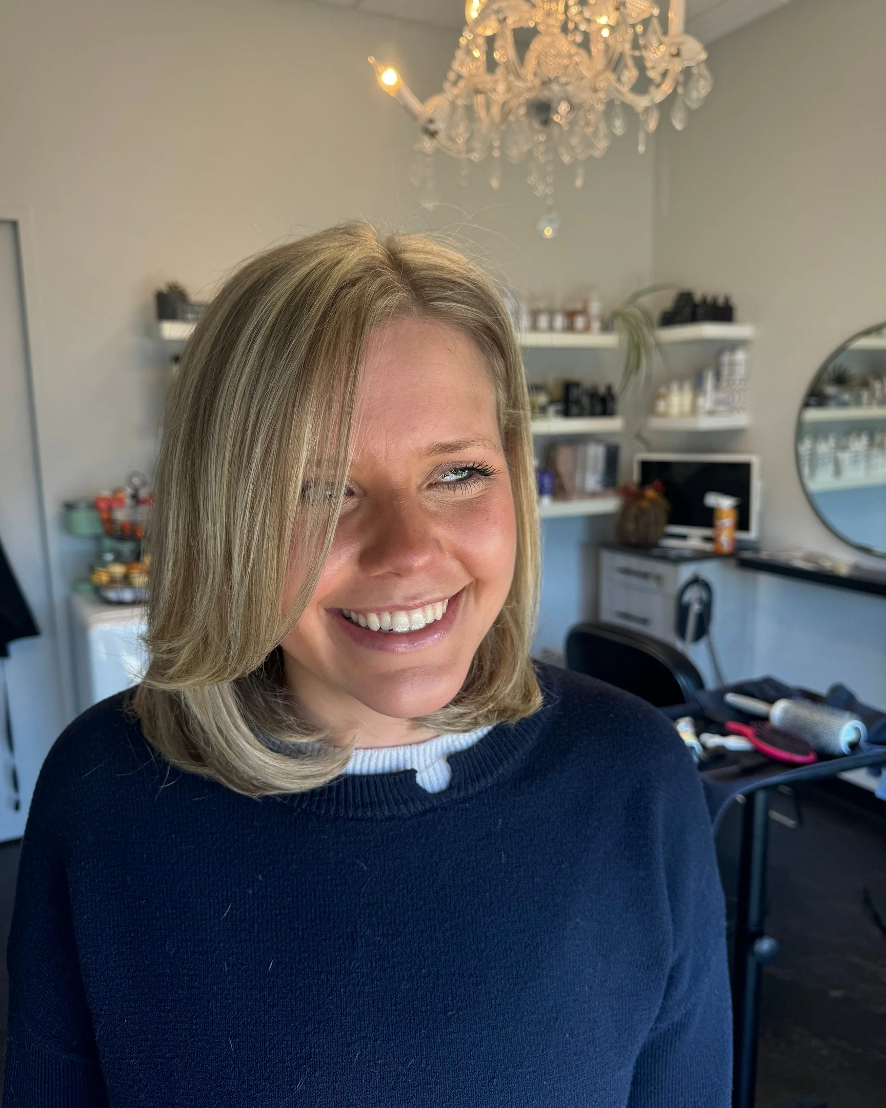 A woman with shoulder-length blonde hair smiling in a salon or makeup studio, with a chandelier hanging above, shelves with products in the background, and beauty tools on a table nearby.