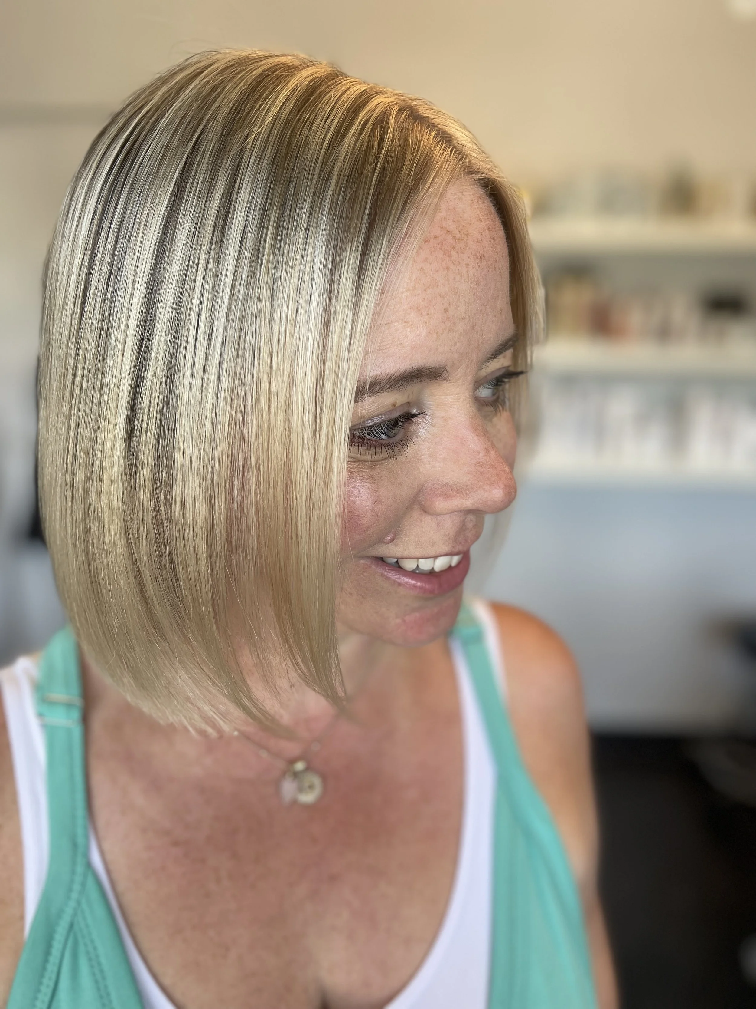 A woman with shoulder-length blonde hair styled in a sleek bob, smiling slightly, wearing a turquoise and white tank top, and a necklace with a small pendant in a bright room.