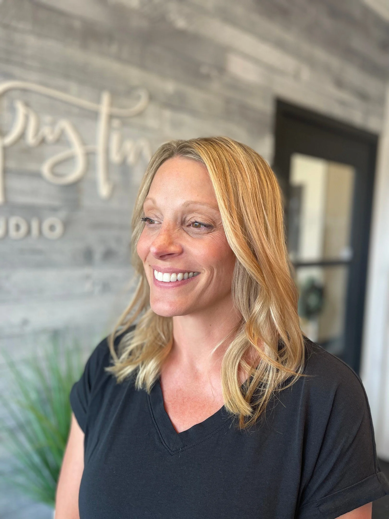 A smiling woman with blonde hair in a black shirt standing inside a studio with a gray wood-paneled wall and a sign that reads 'Insta'.