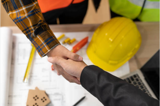 Two people shaking hands over a construction site desk with a yellow safety helmet, blueprints, ruler, and pens.