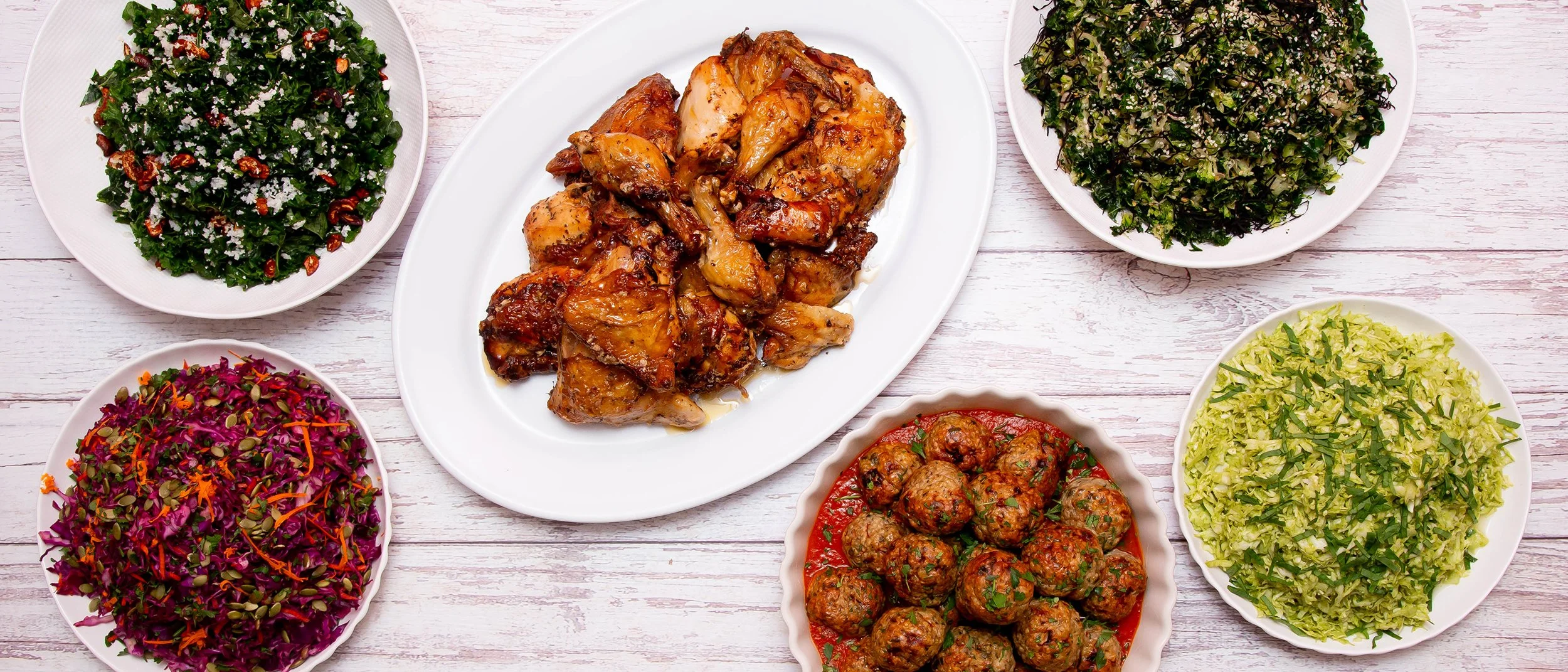 Assorted Middle Eastern dishes including grilled chicken, tabbouleh, chopped kale salad, cabbage salad, chopped parsley salad, and pomegranate and pecan salad on white plates.