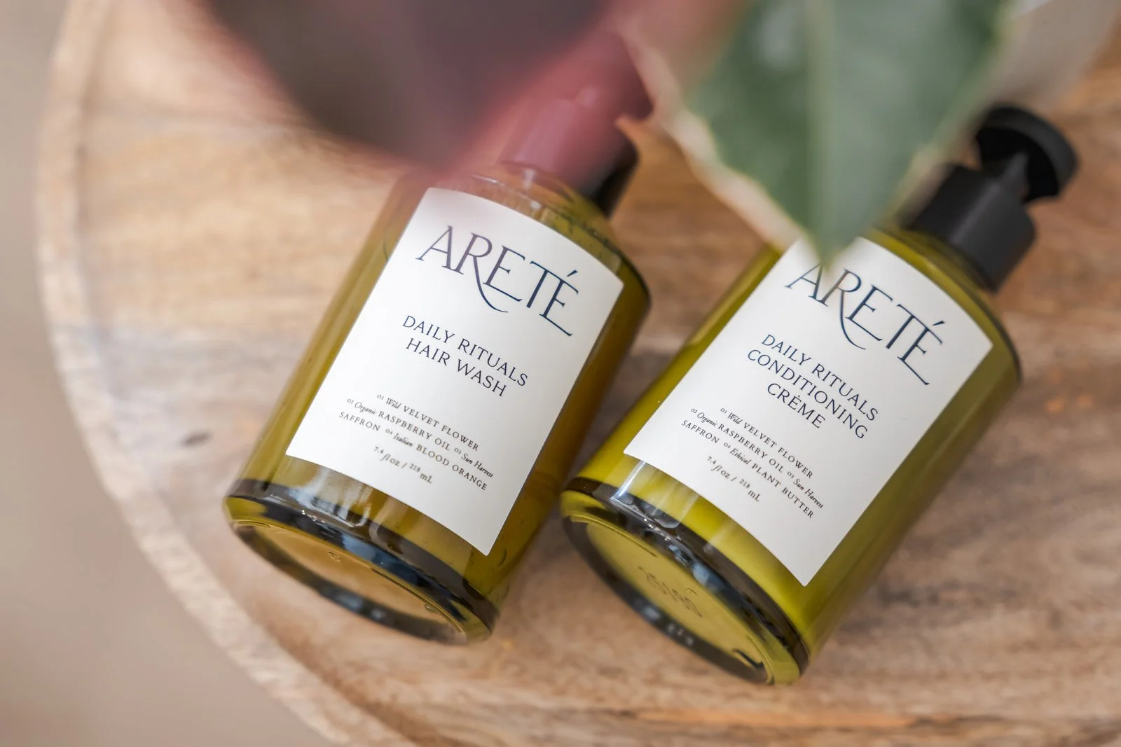 Two bottles of Areté hair and conditioning products on a wooden surface, one labeled 'Daily Rituals Hair Wash' and the other 'Conditioning Crème', with botanical ingredients listed on the labels.