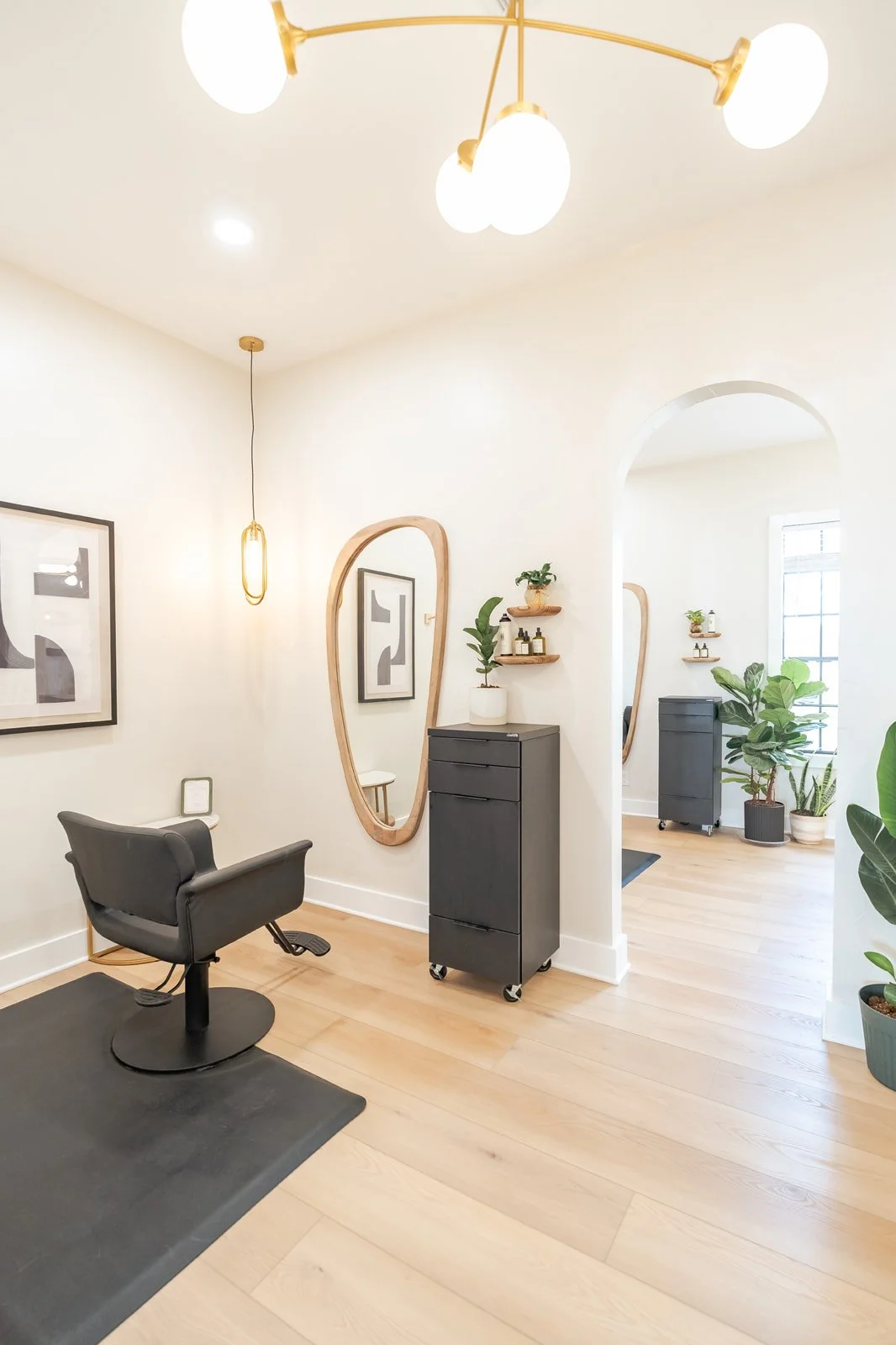 Modern hair salon with black styling chair, large wooden hanging mirror, black rolling storage cart, and potted plants in a bright space with white walls and wood flooring.