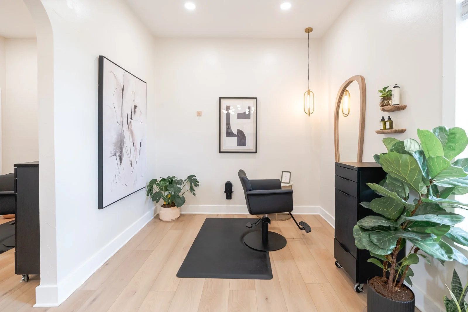 Minimalist luxury hair salon with black station and chair, abstract wall art, potted plants, and warm pendant lighting.
