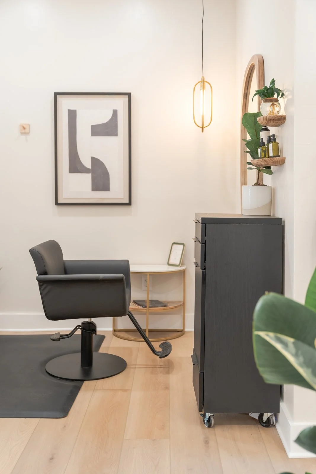 Hair styling station in a luxury salon with a black storage cabinet, plant, clean products on a shelf, and a hanging light fixture.