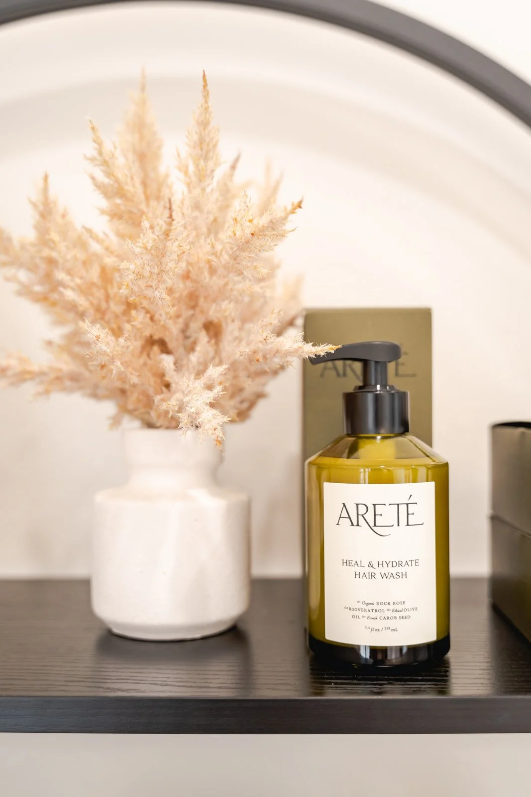 A bottle of Aretė Heal & Hydrate Hair Wash next to a white vase with pink dried pampas grass on a black shelf.