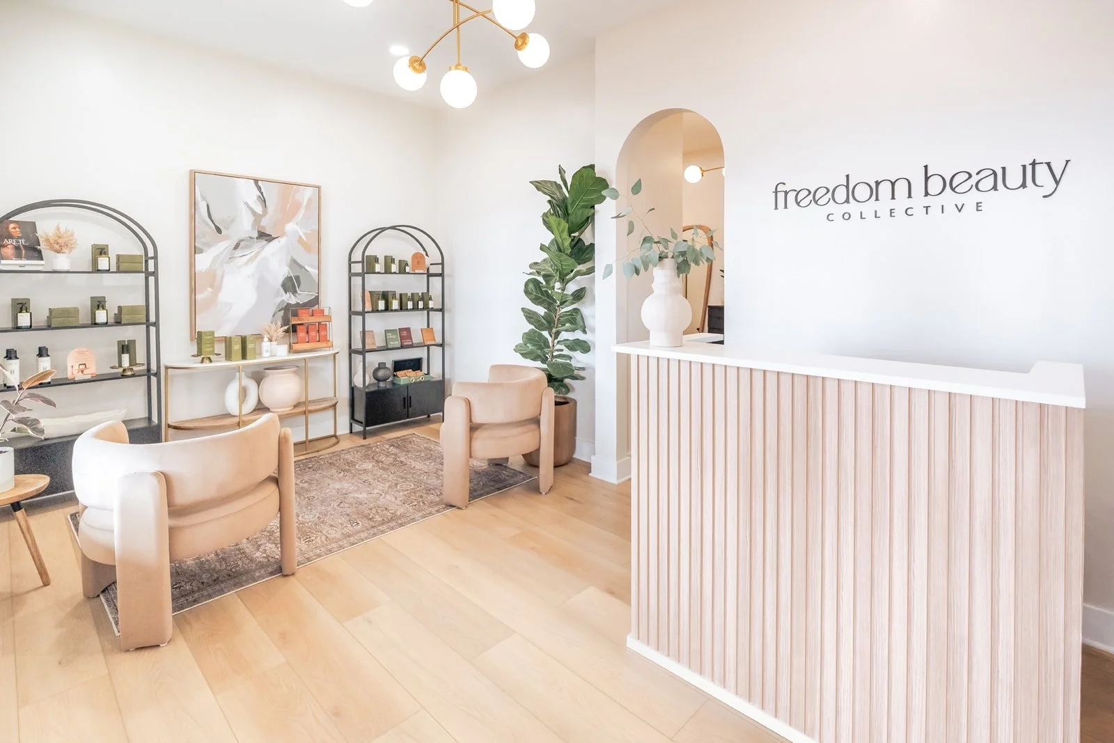 Interior of a modern, low-tox hair salon with a reception desk, beige chairs, metal shelving displaying clean beauty products, a large abstract painting, and greenery with a sign that says "freedom beauty COLLECTIVE."