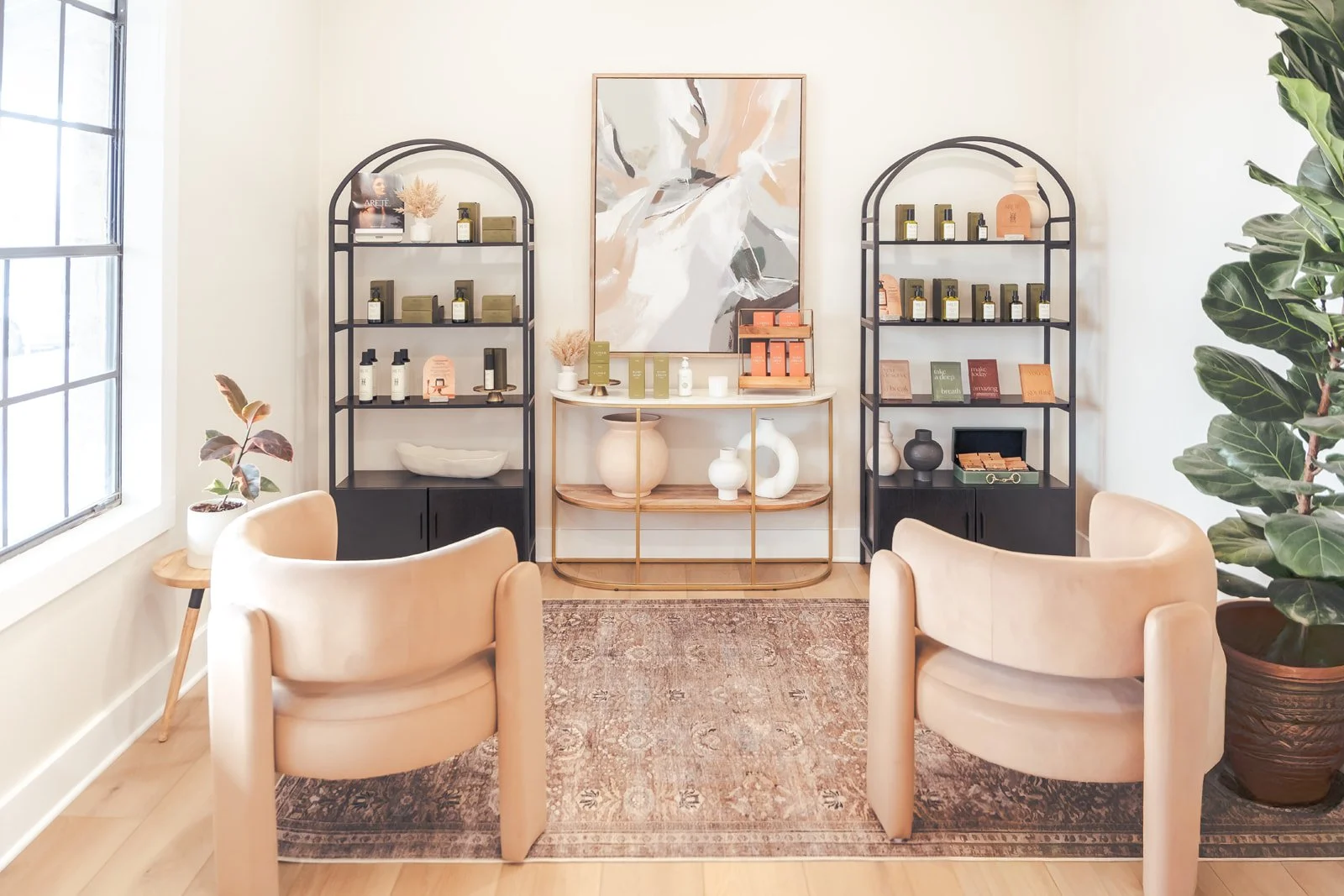 A cozy, modern retail space with two beige armchairs facing a display wall. The wall has black metal shelves filled with products, with a framed abstract art piece above a gold and glass console table displaying vases and decor items. A large potted 