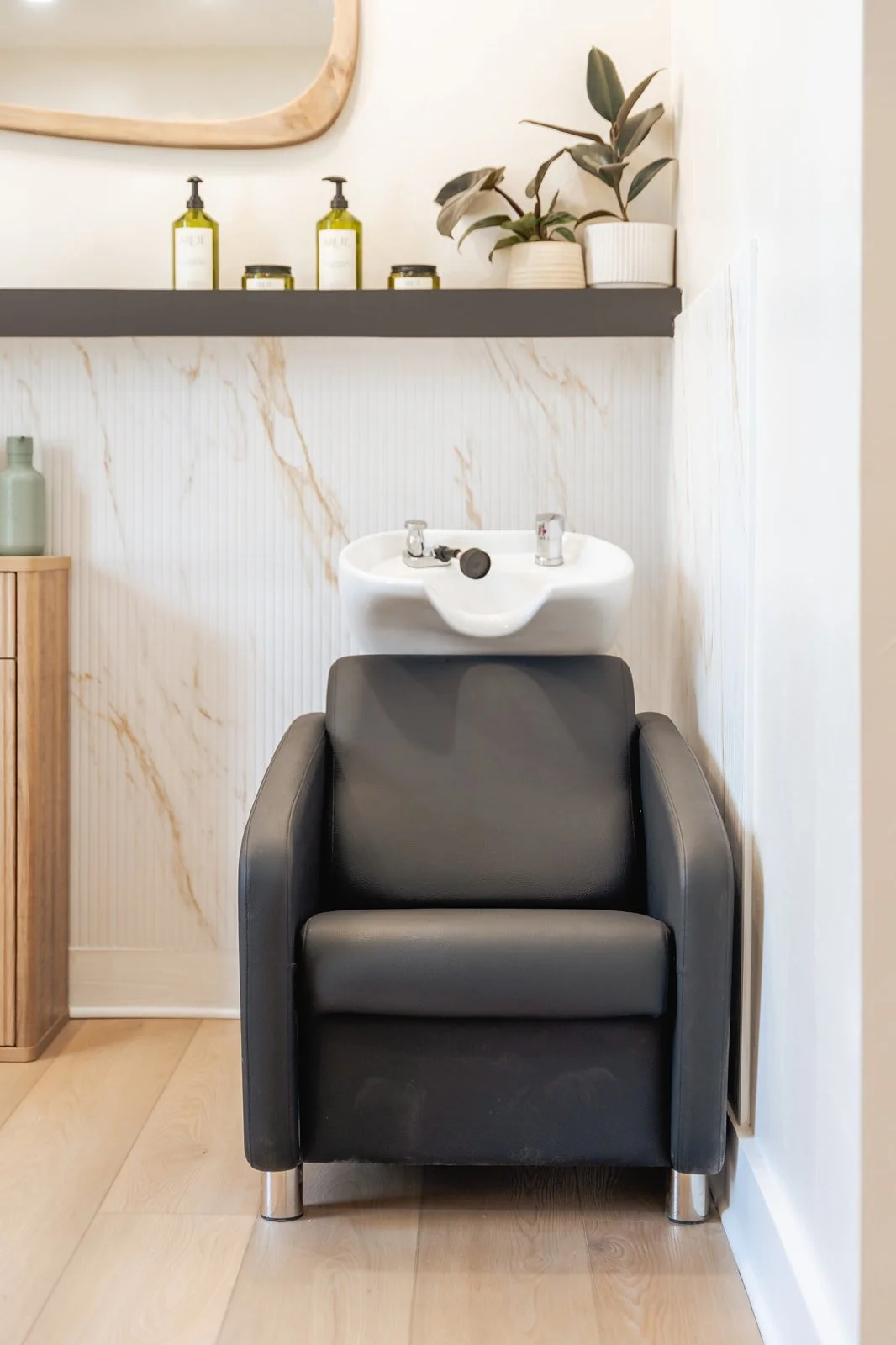 A black shampoo chair with a white ceramic sink attached behind it, set against a wall with white and beige marble-like paneling. A shelf above holds green bottles, candles, and a plant in a white pot.