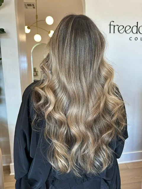 Back view of a woman with long, wavy blonde hair in a salon.