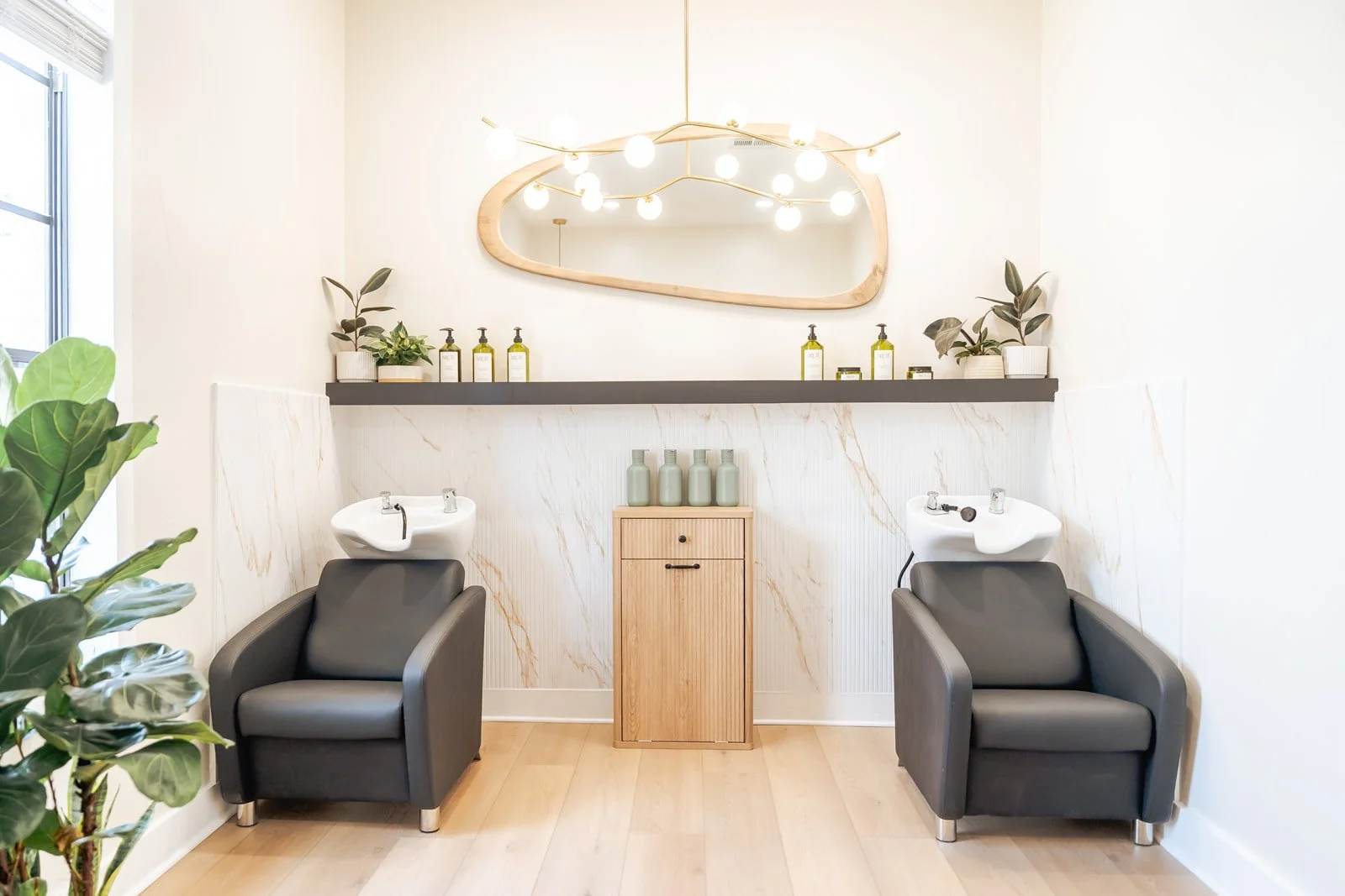 Hair salon with two black salon chairs and white shampoo bowls, wooden cabinet in the center, wall shelf with green shampoo bottles and potted plants, a large mirror with wooden frame, and a plant in the foreground.