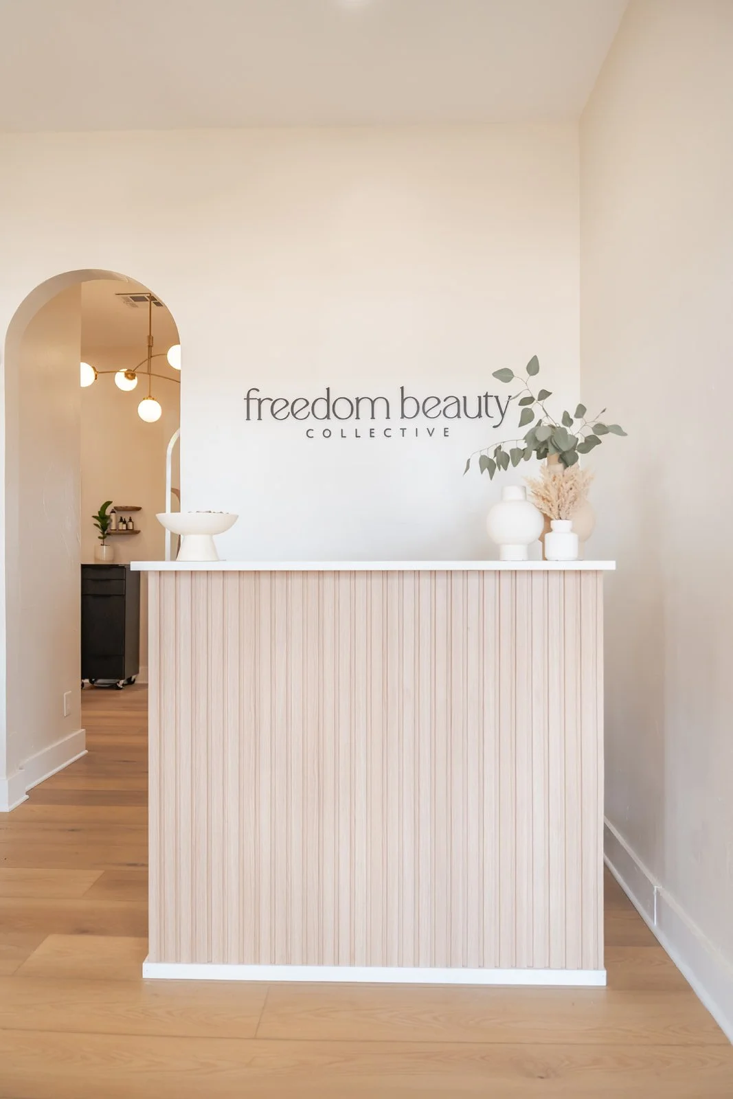 Reception desk with minimalist decor at Freedom Beauty Collective, a low-tox luxury hair salon featuring white and beige tones, vases with eucalyptus, and a sign on the wall.
