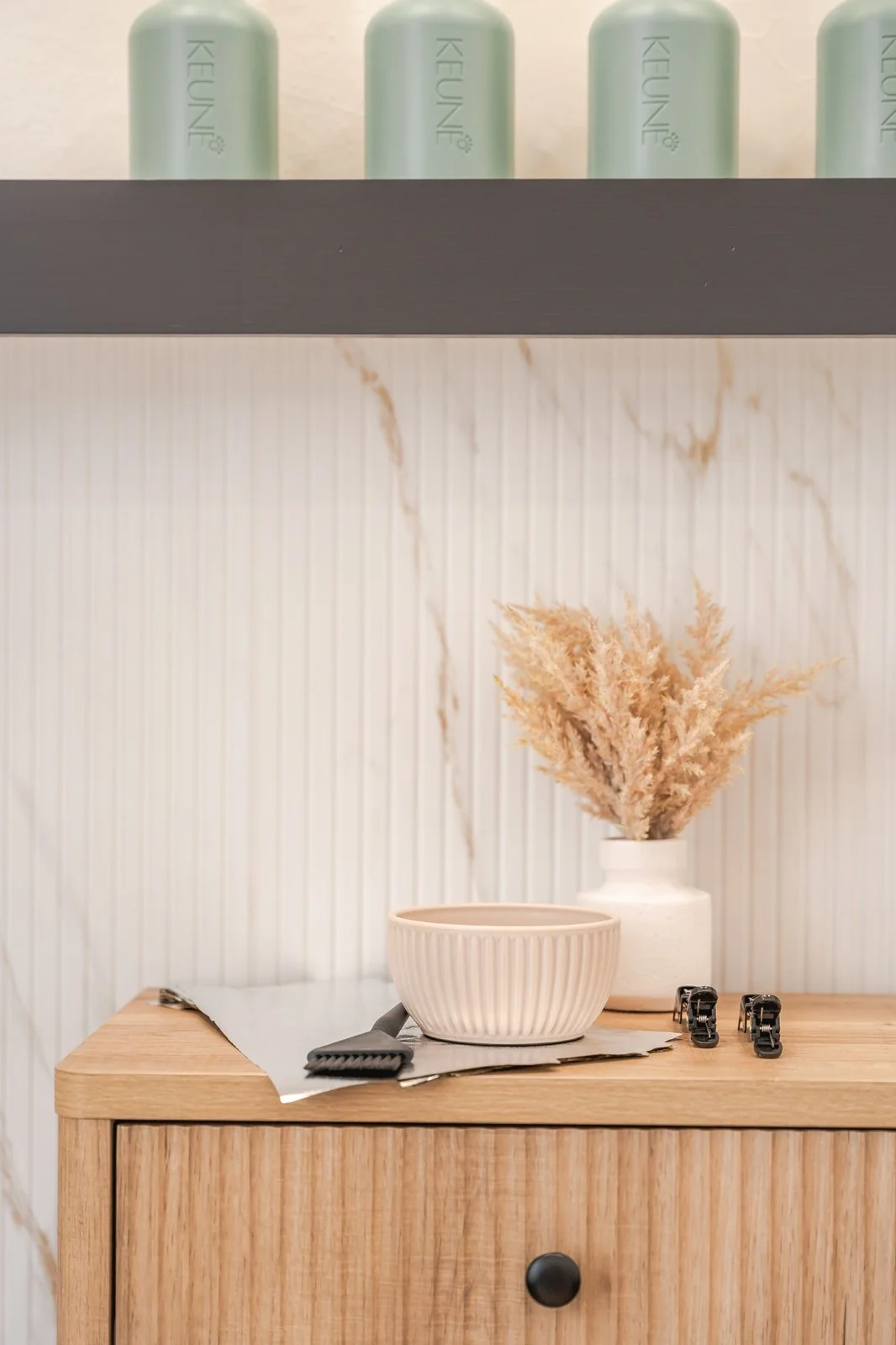 A wooden cabinet with a beige ribbed bowl, a small white vase with dried pink pampas grass, a hairbrush, and two hair clips on top. The background features a white, marbled wall with vertical ridges.