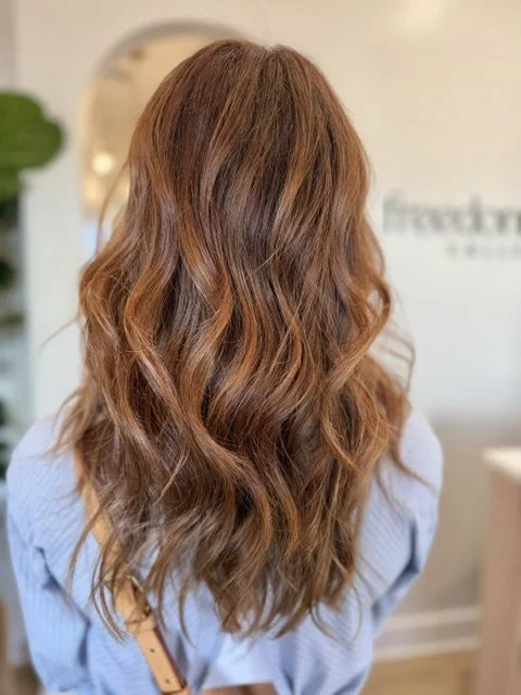 Back view of a woman with long, wavy, reddish-brown hair inside a hair salon.