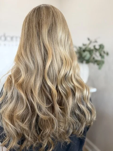 Back of a woman with long, blonde, wavy hair standing in a luxury hair salon. 