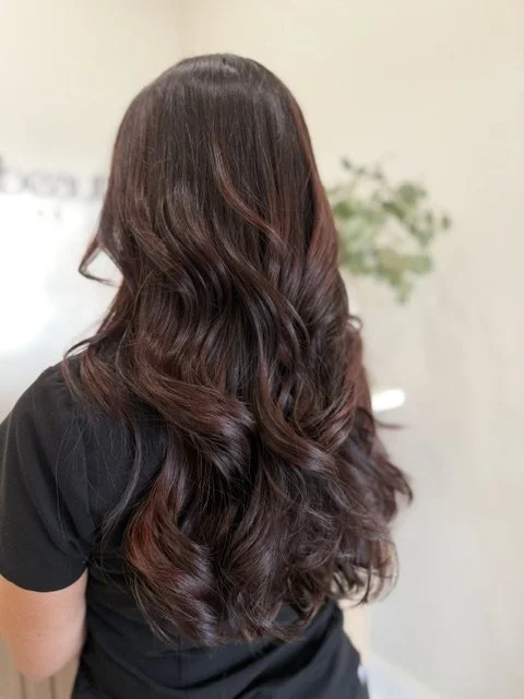 Back view of a woman with long, wavy, dark brown hair with red highlights in a luxury hair salon.