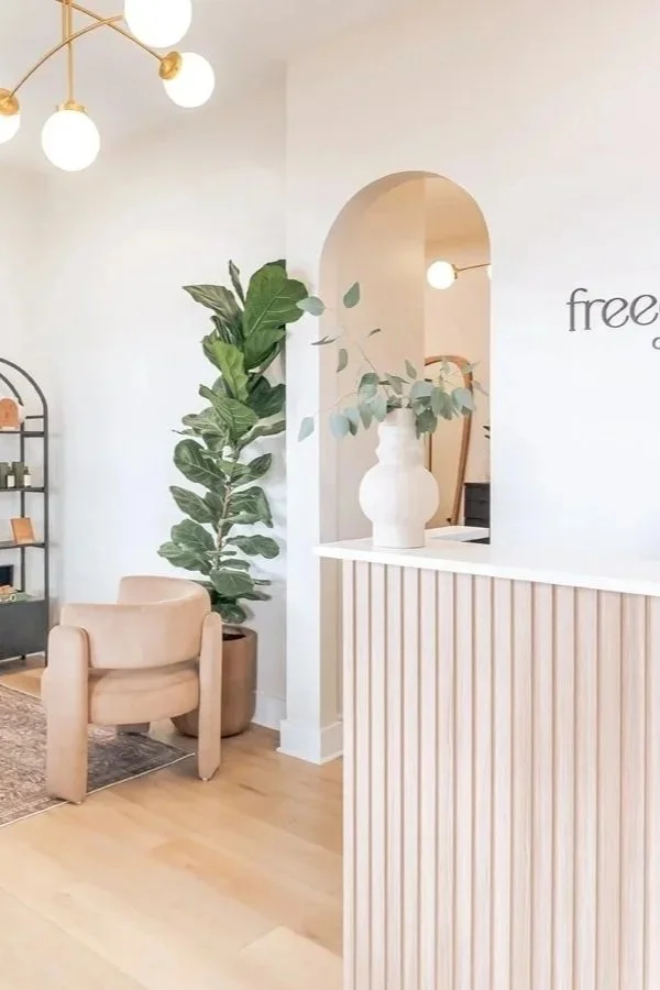 Interior of a modern, minimalist room with a beige armchair, a large potted fiddle leaf fig plant, a white ceramic vase with greenery, and a wooden slatted divider. There are round globe lights and an arched opening in the background.