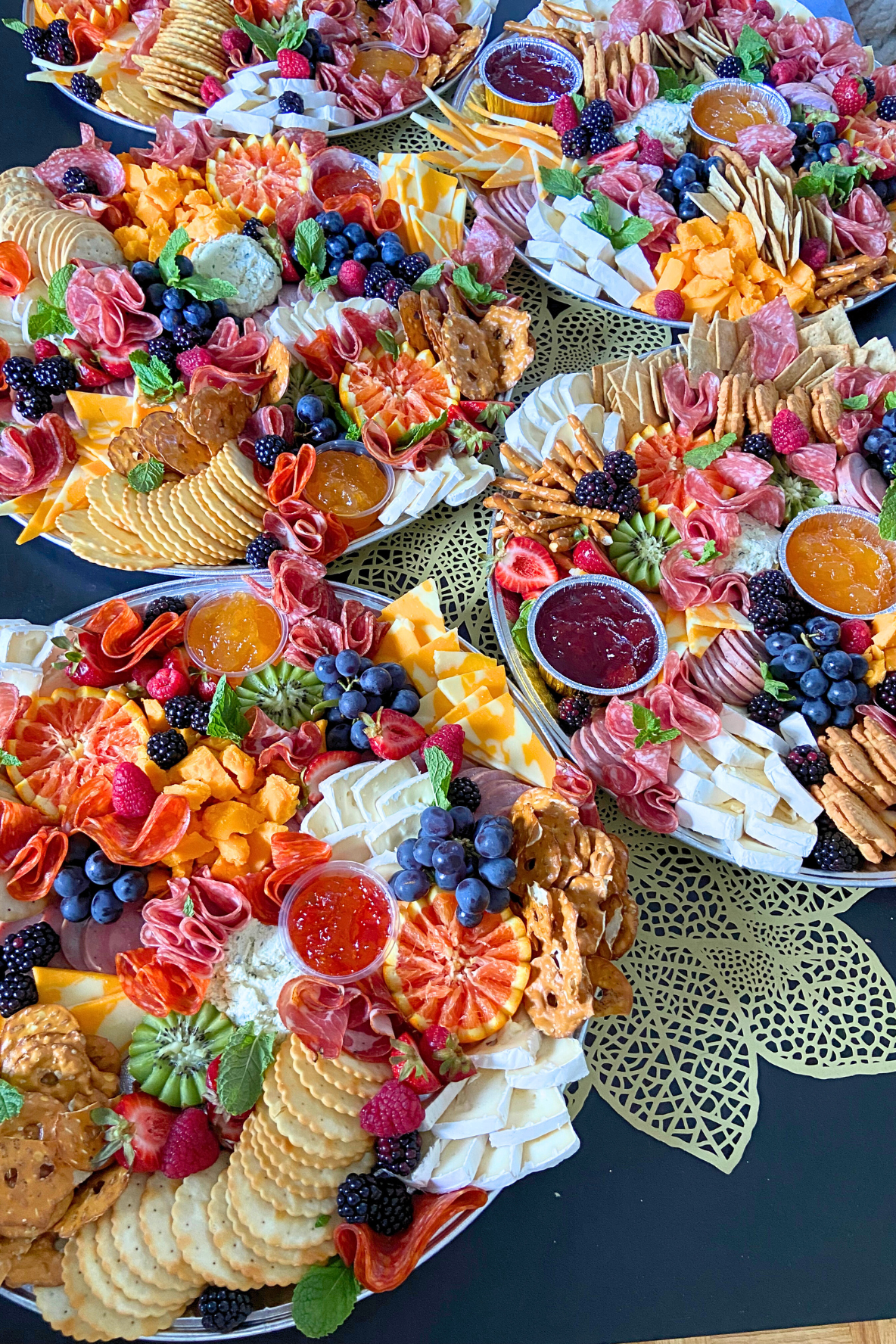 Close-up of several large trays filled with assorted cheese, fresh fruits, crackers, and small bowls of jams and spreads, arranged for a buffet