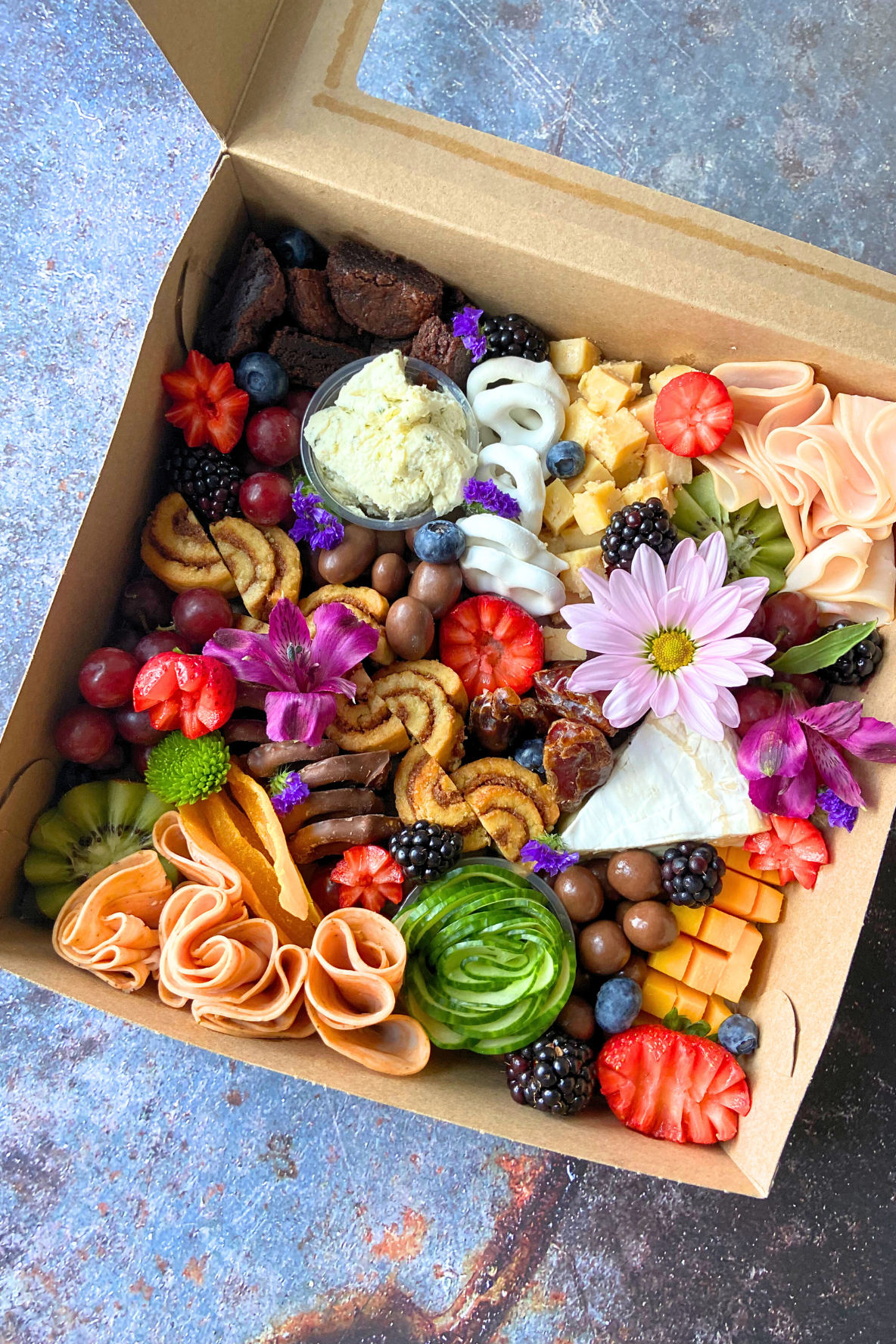 A box filled with a colorful assortment of cheeses, fruits, chocolates, and flowers.