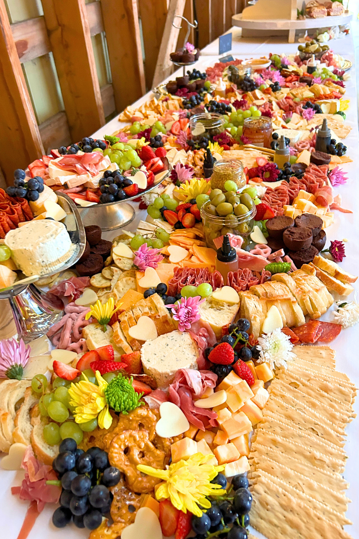 A long table filled with a variety of cheeses, sliced meats, fresh grapes, strawberries, blueberries, crackers, and flowers.