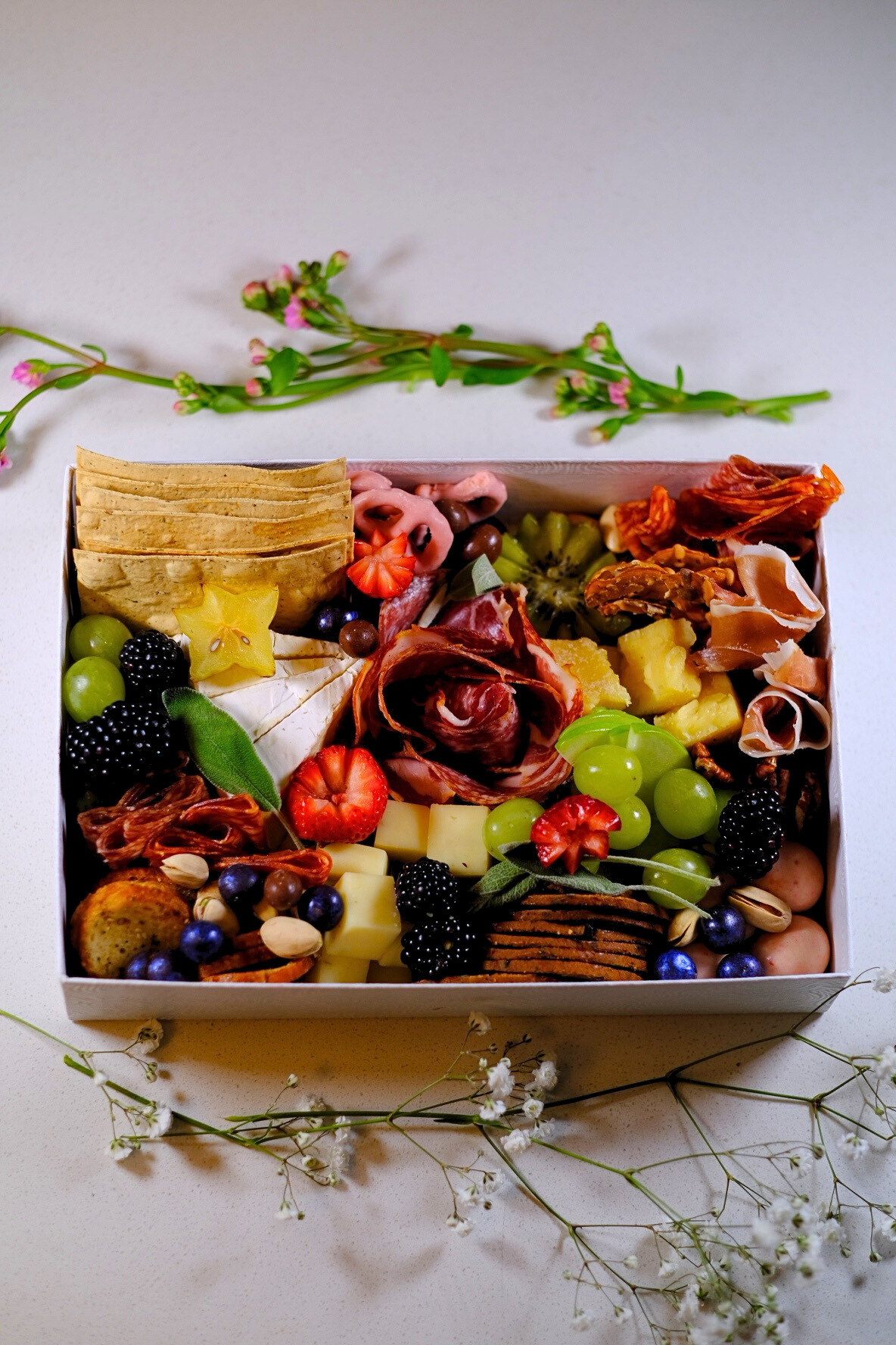 A rectangular cheese and charcuterie board with grapes, blackberries, strawberries, sliced kiwis, various cheeses, crackers, prosciutto, and other cured meats, surrounded by sprigs of small white flowers.