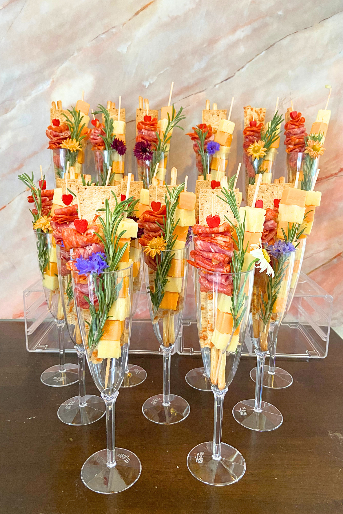 Appetizer served in tall glasses with multiple layers of cheese, cured meats, crackers, and garnished with sprigs of herbs and small edible flowers.