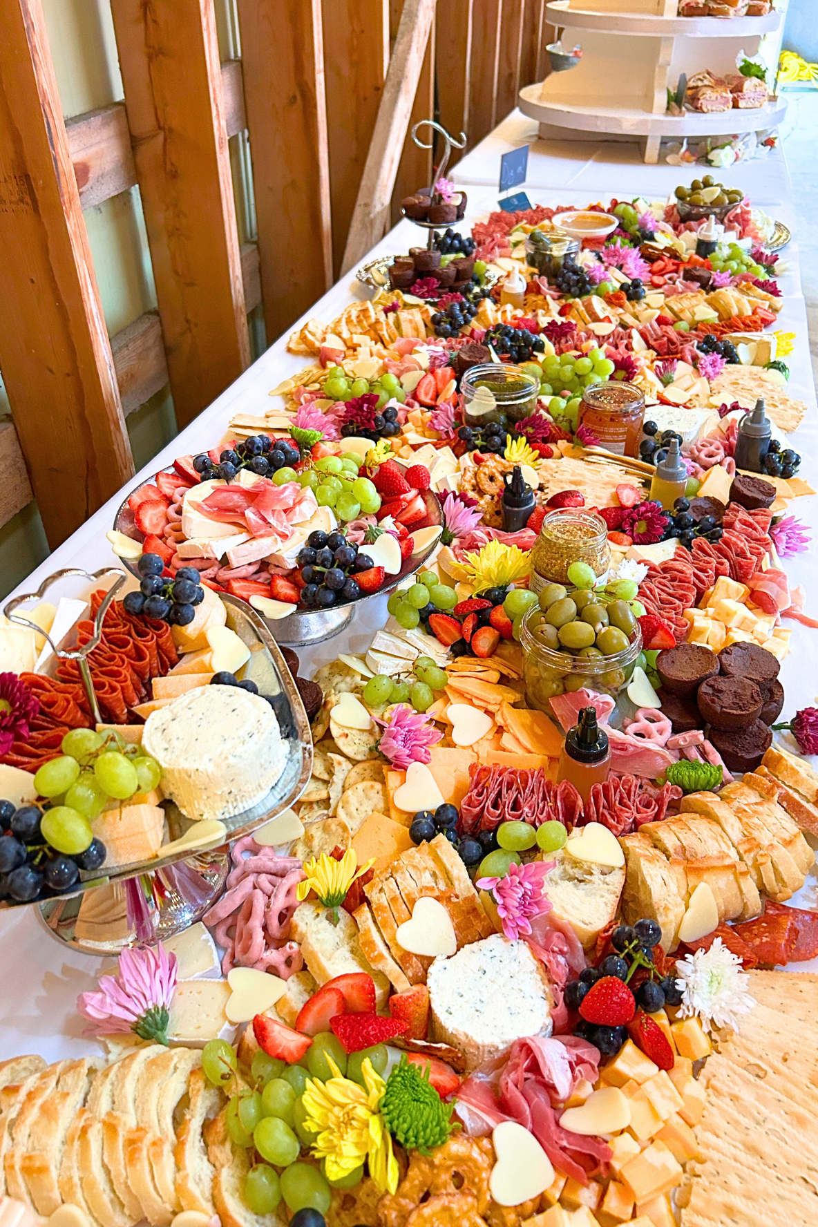 An elaborate spread of various cheeses, meats, fresh fruits, flowers, and condiments on a long table, set for a buffet.