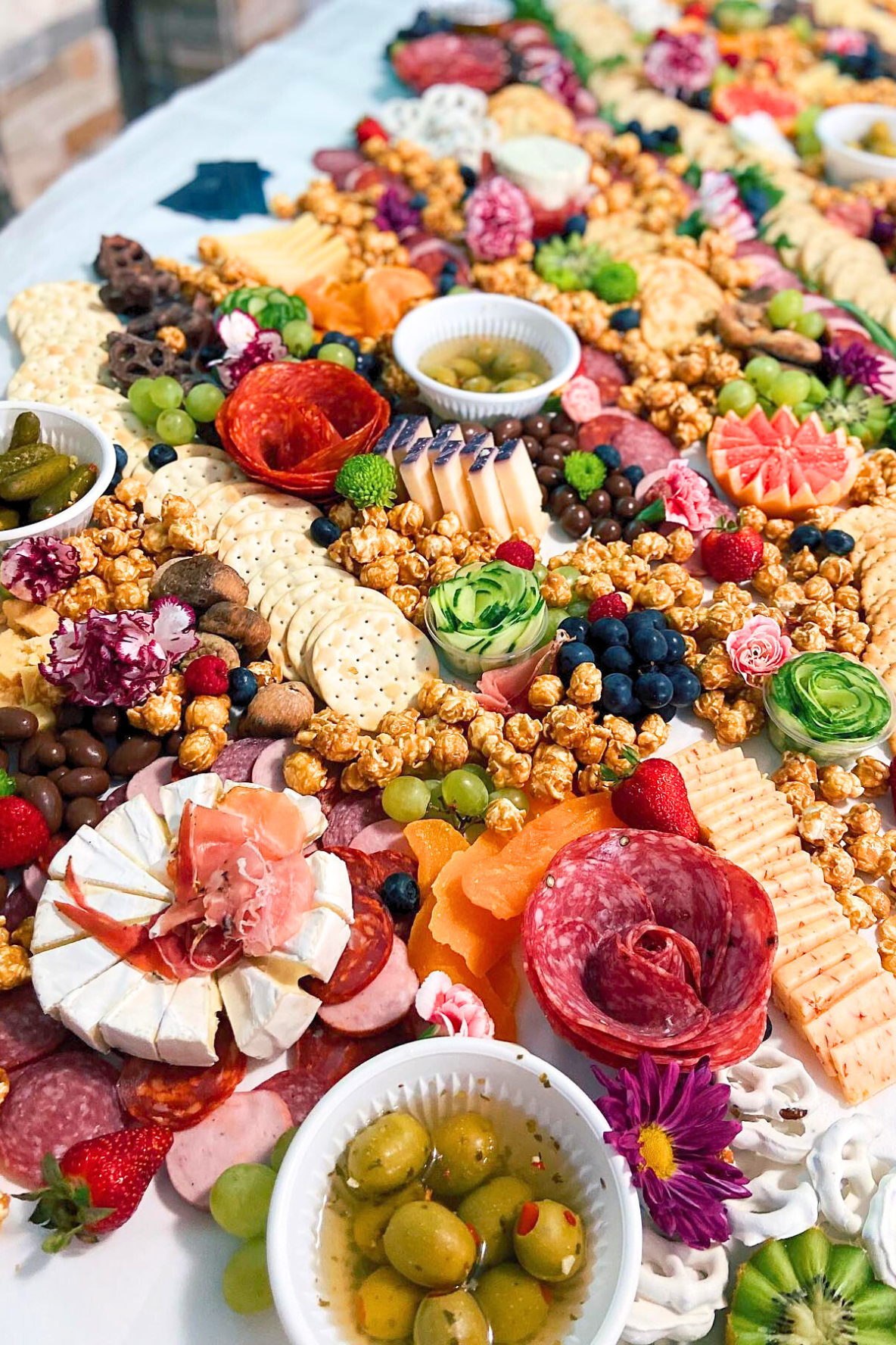 A large charcuterie board with an assortment of cheeses, cured meats, crackers, grapes, strawberries, olives, and flowers.