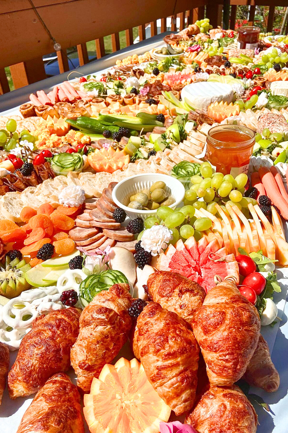 A large outdoor table filled with an assortment of breakfast or brunch foods including croissants, various cheeses, meats, fresh fruits like grapes, watermelon, strawberries, and oranges, along with jars of jam, and vegetables. The table is set on a 