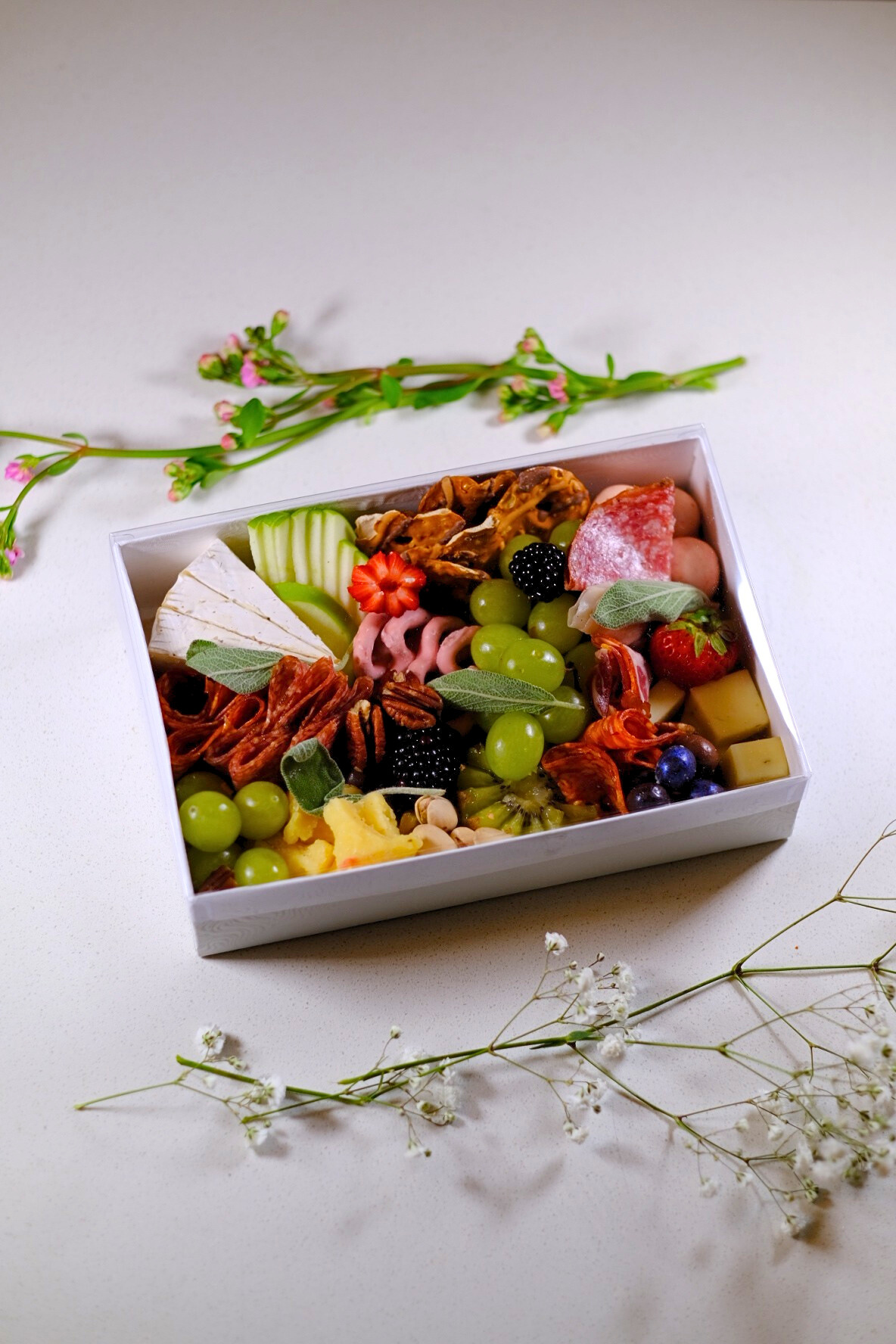 A box of assorted cheeses, fruits, nuts, and snacks on a white surface with sprigs of white and pink flowers.