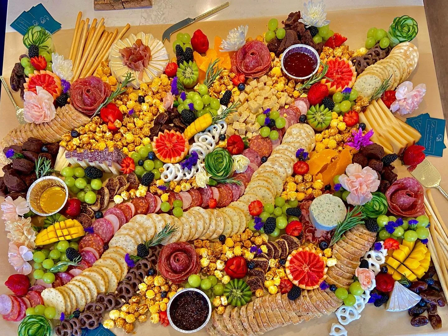 A large cheese and charcuterie board decorated with various vegetables, fruits, crackers, meats, cheeses, popcorn, and edible flowers.