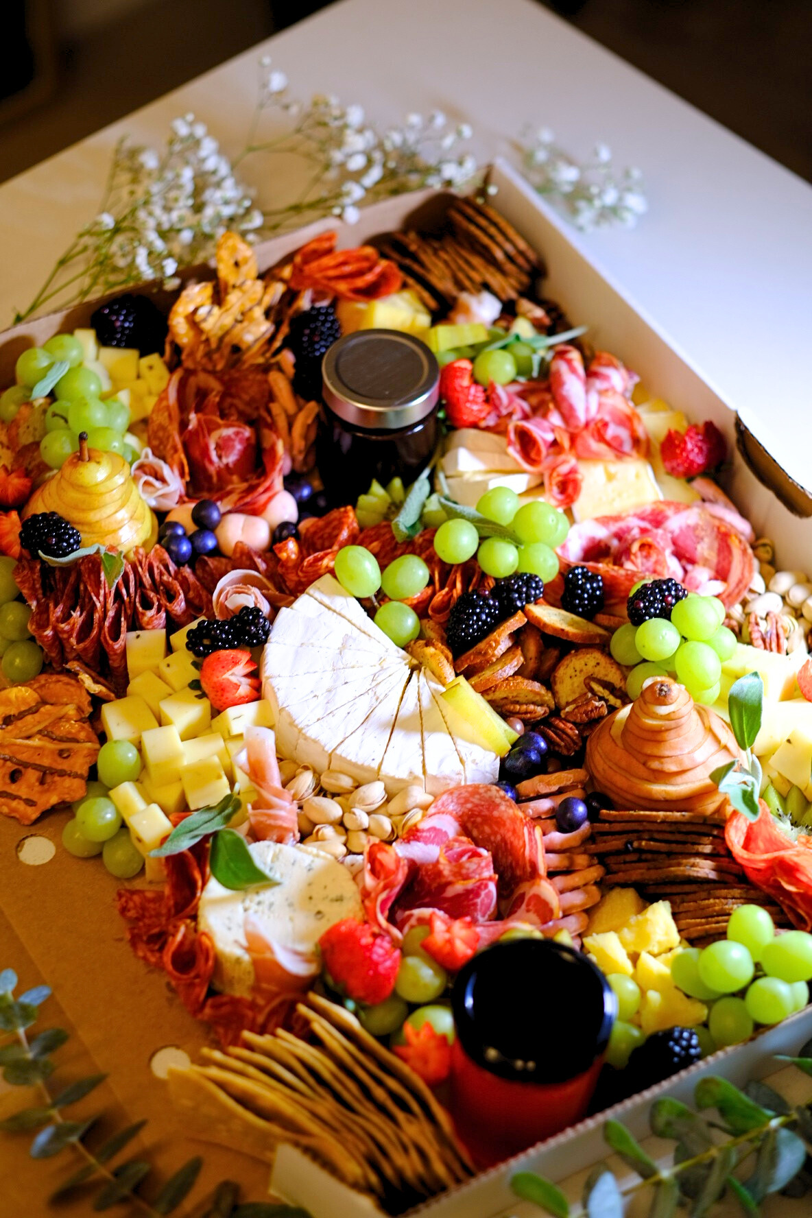 A large charcuterie and cheese platter with grapes, strawberries, dried fruits, nuts, various cheeses, crackers, and small jars, decorated with sprigs of greenery and white flowers.