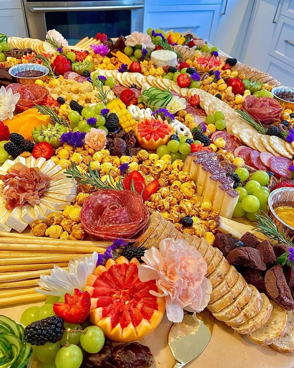 A colorful, elaborate charcuterie and cheese spread with fruits, crackers, flowers, and various snacks on a large table.