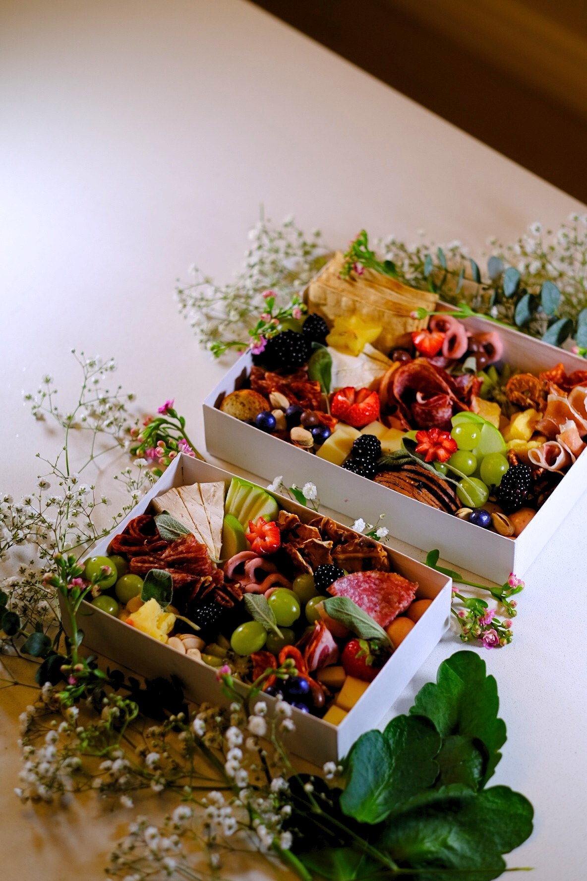 Two boxes of assorted charcuterie and cheese, garnished with fresh grapes, berries, and herbs, placed on a decorated table.