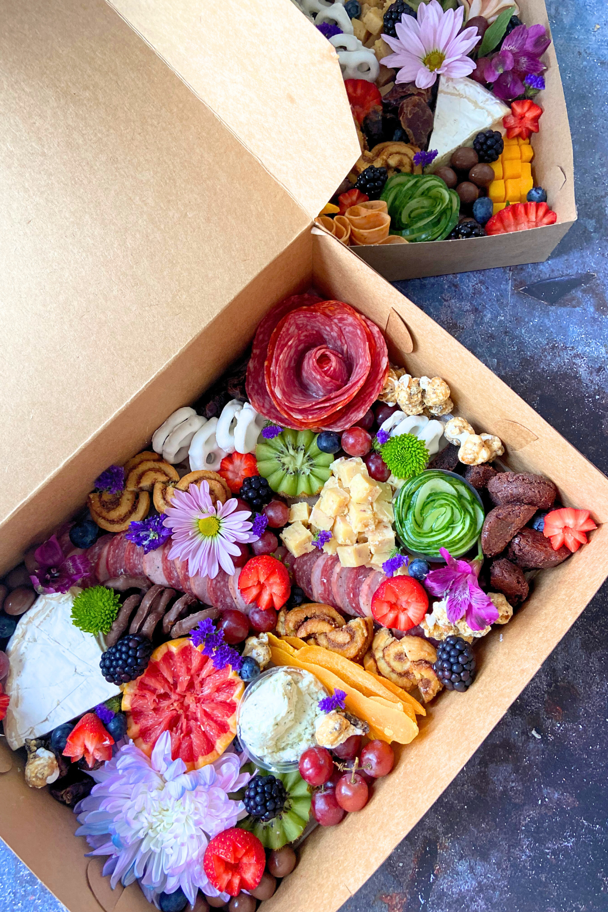 Two large cheese and charcuterie boards with various cheeses, cured meats, fresh and dried fruits, nuts, and edible flowers.