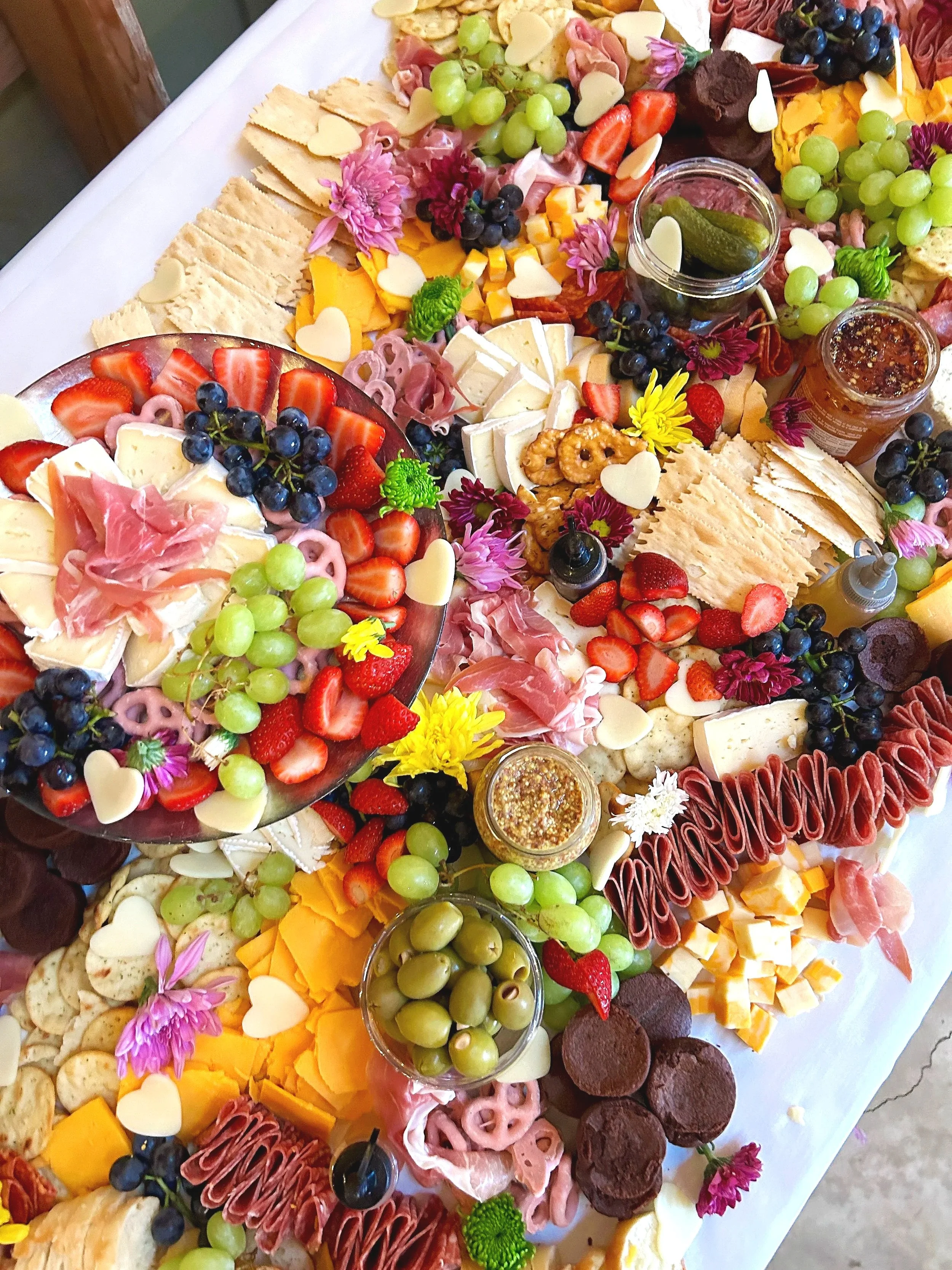 A cheese and fruit platter with grapes, strawberries, olives, meats, crackers, and condiments on a white tablecloth, decorated with edible flowers.