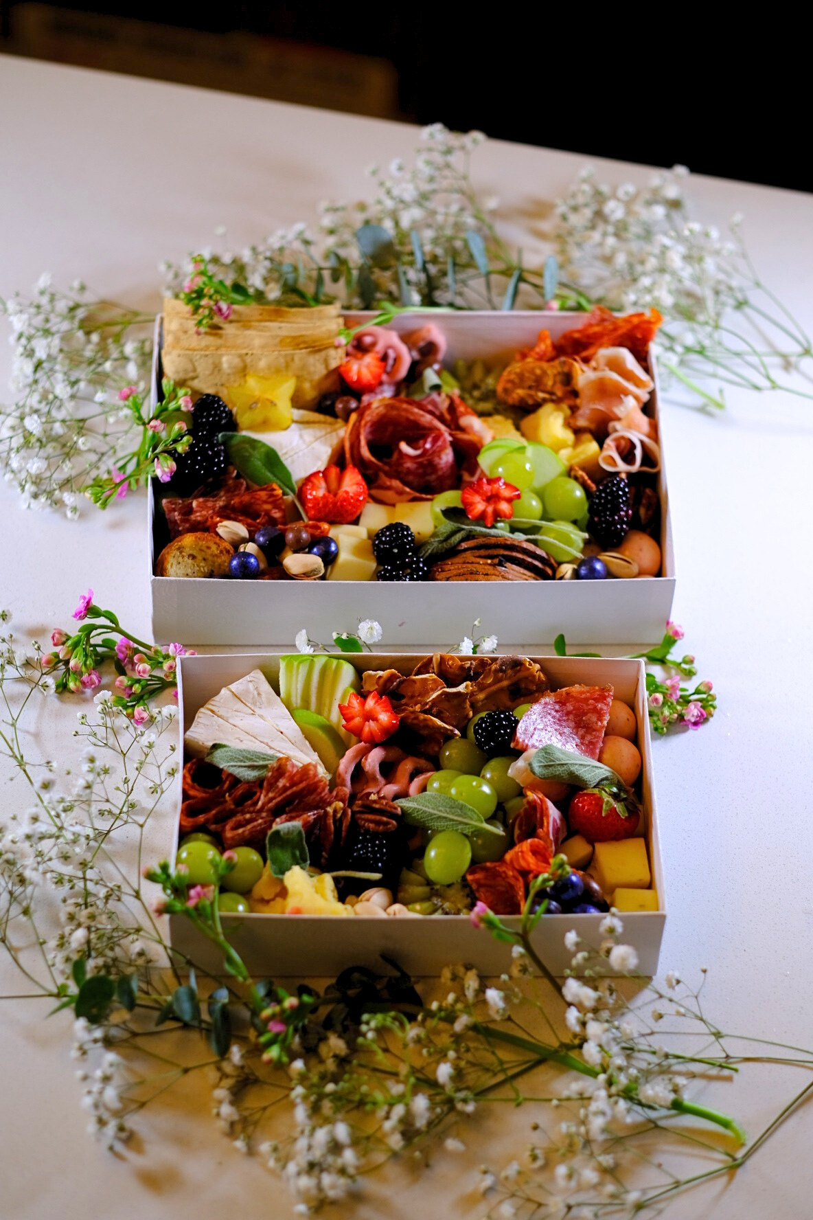 Two open boxes filled with assorted cheeses, cured meats, grapes, strawberries, blackberries, nuts, and crackers, decorated with white and pink flowers on a white table.