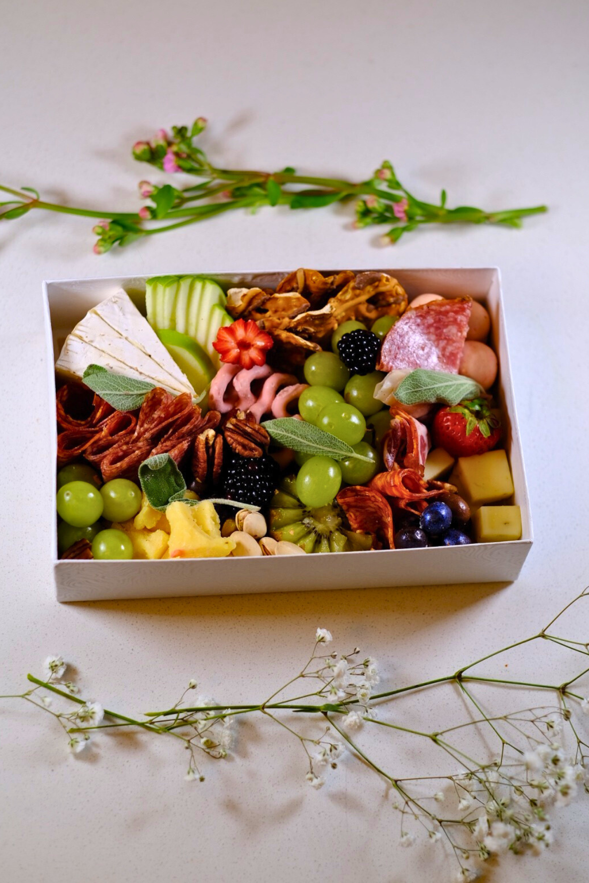 A box of assorted cheeses, grapes, nuts, berries, and cured meats, surrounded by sprigs of flowers on a white surface.